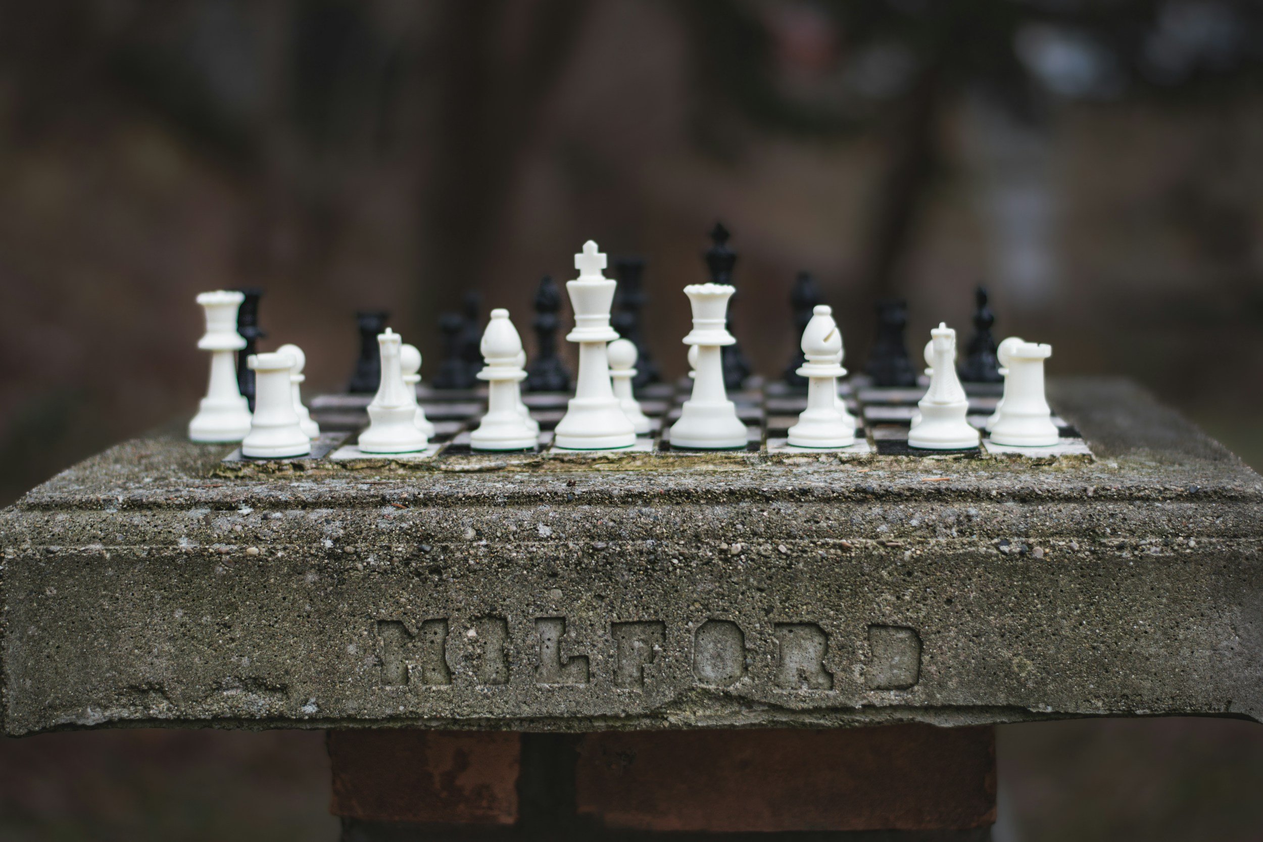 A picture of a concrete chess table, likely in an outdoor area, with chess pieces in their starting places. White chess pieces are closest to the camera and black are fuzzy in the background. The concrete table has Milford stamped into it.