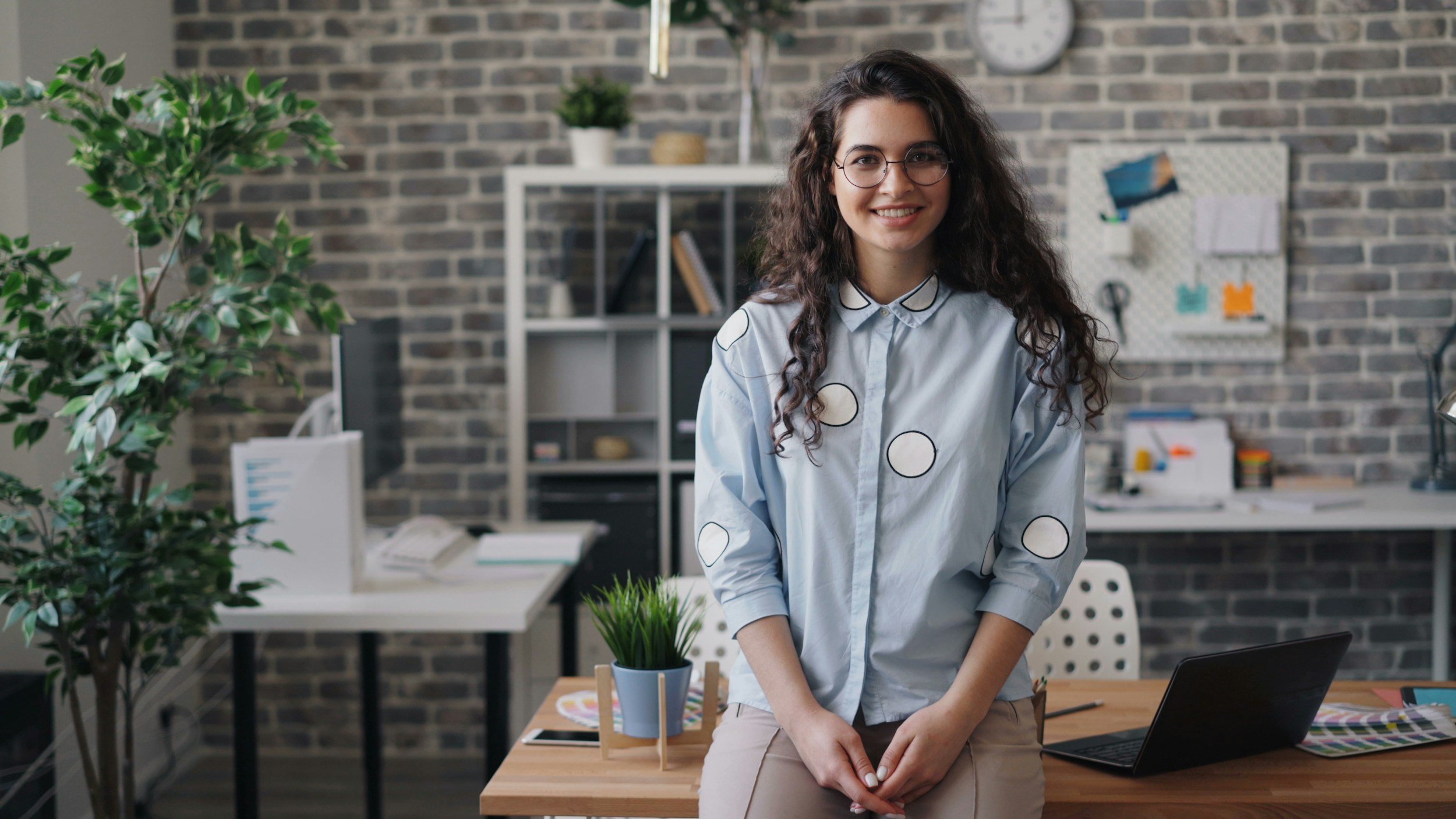 Young white women with long dark brown curly hair, wearing a light blue button up shirt with white polka dots and tan pants, sitting on a work desk with a laptop. She is facing the camera and smiling, with thin rimmed glasses on, hands in her lap.