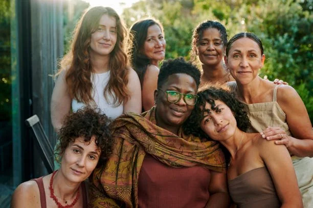 Several women of various ages and ethnicities standing together, looking at the camera contently. There are trees in the background that are blurred out.