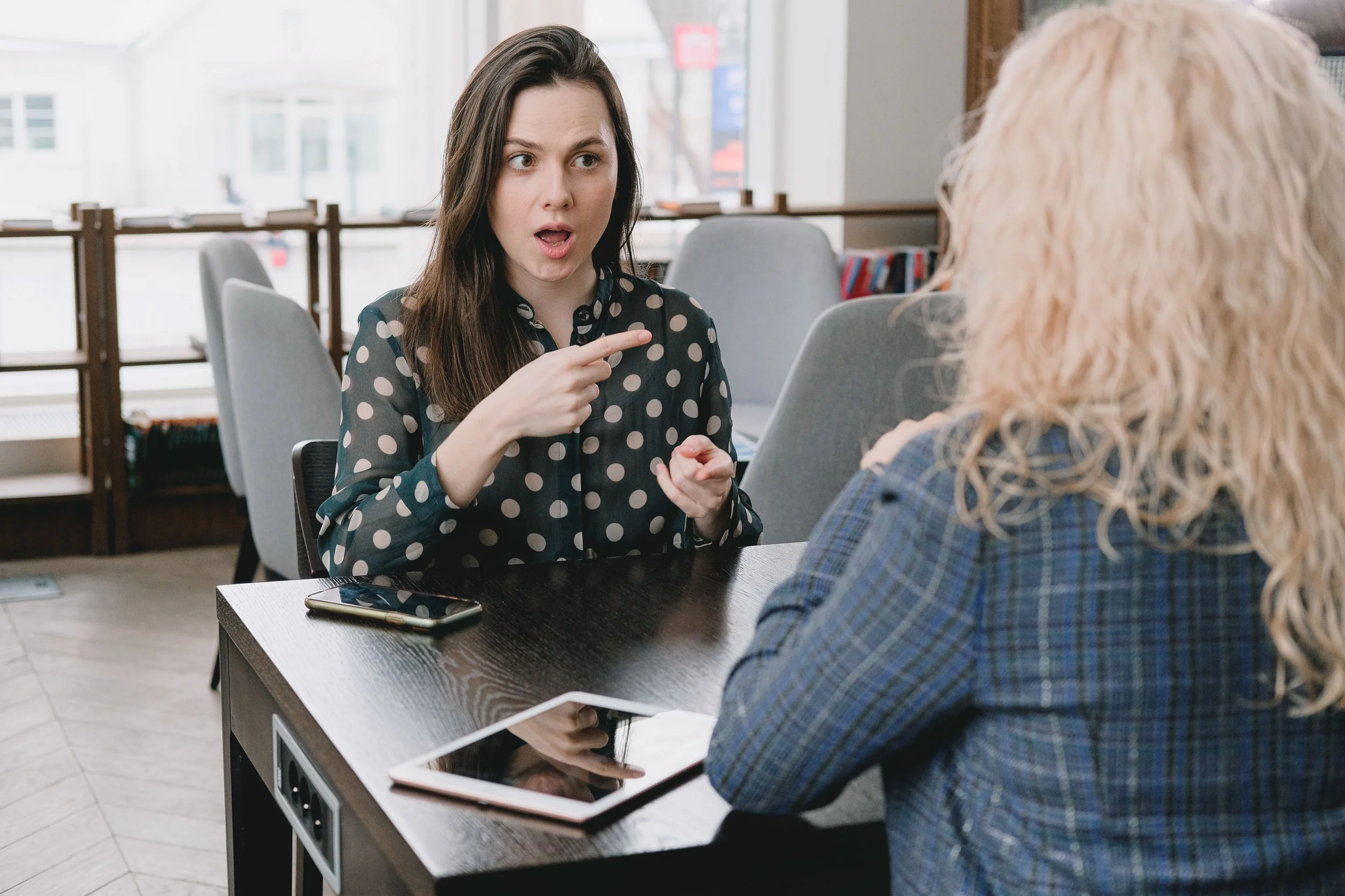 Two women talking across a table. One is wearing a black sheer blouse with cream polka dots. She has long, straight brown hair, her hands in front of her, pointing her fingers while talking. The other is blonde with a blue blazer, facing away .
