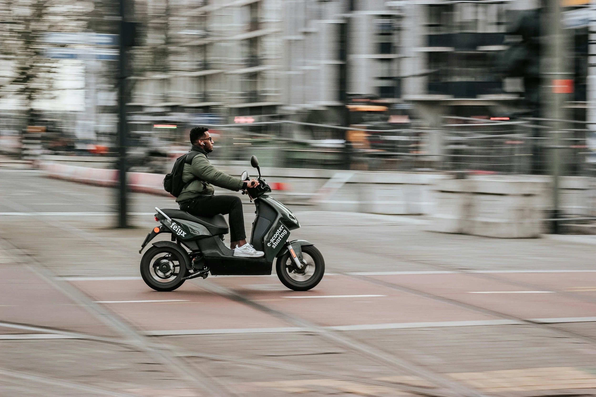 Brown skinned man with short, cropped hair, black framed glasses, and ear buds in is wearing a green jacket, black pants, and white shoes with a black backpack. He is riding a gray scooter on a city street. The background is blurred as if moving fast