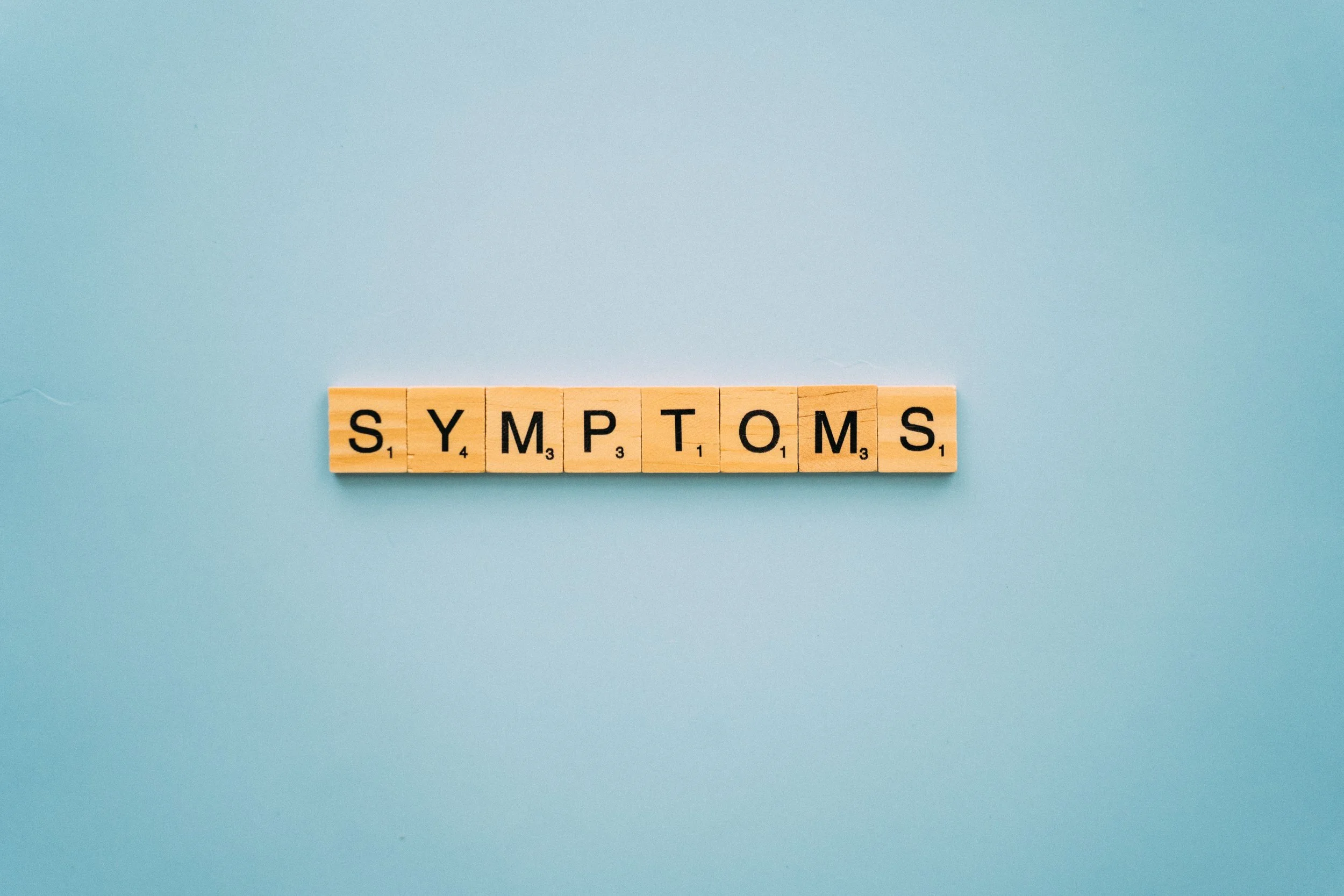 Image of wooden Scrabble tiles side by side that spell out the word 'SYMPTOMS' with a light blue background.