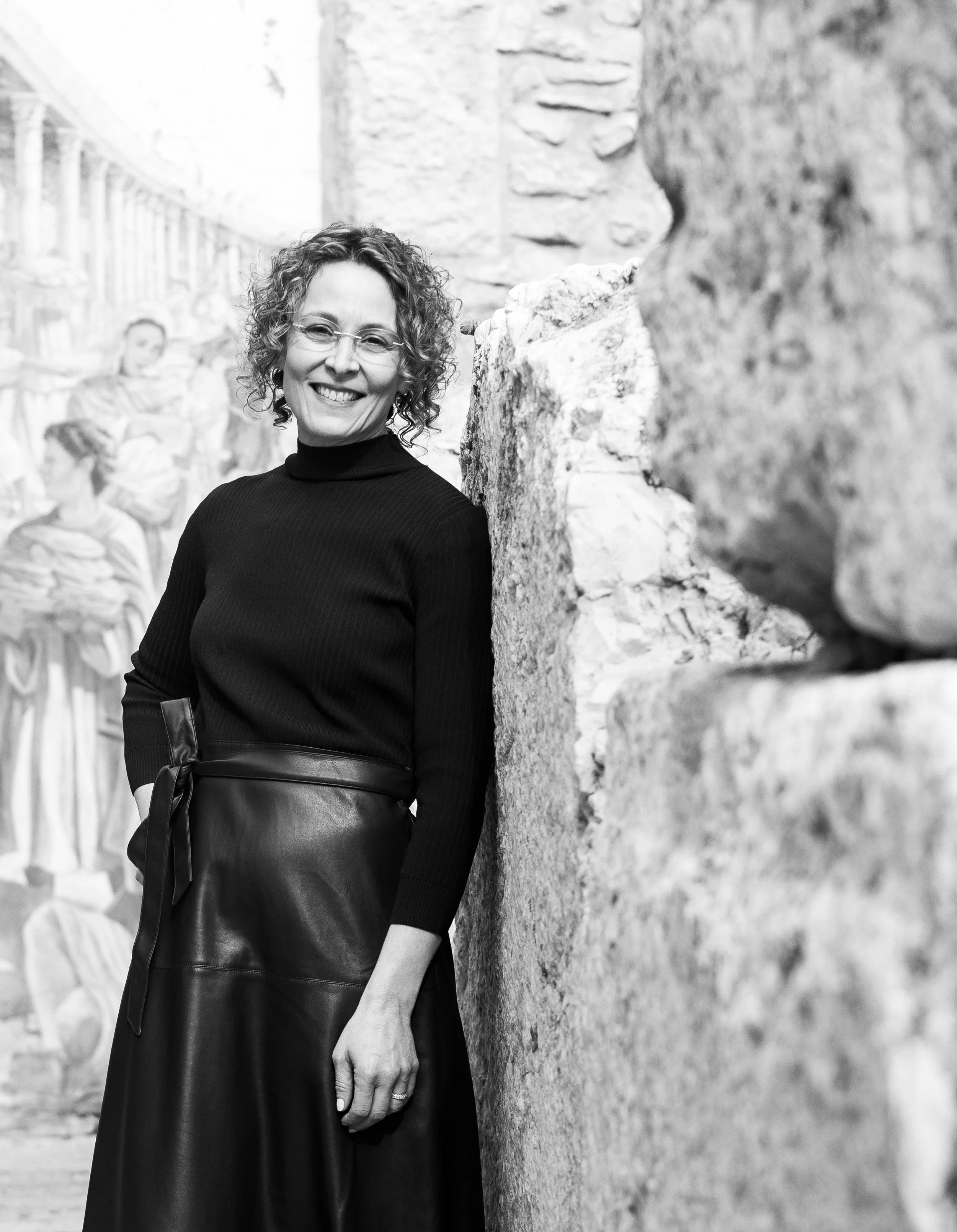 A black and white photograph of Stacey Brickman, a woman with curly hair, glasses, and a bright smile, leaning against a stone wall with a mural or painting of people in the background.