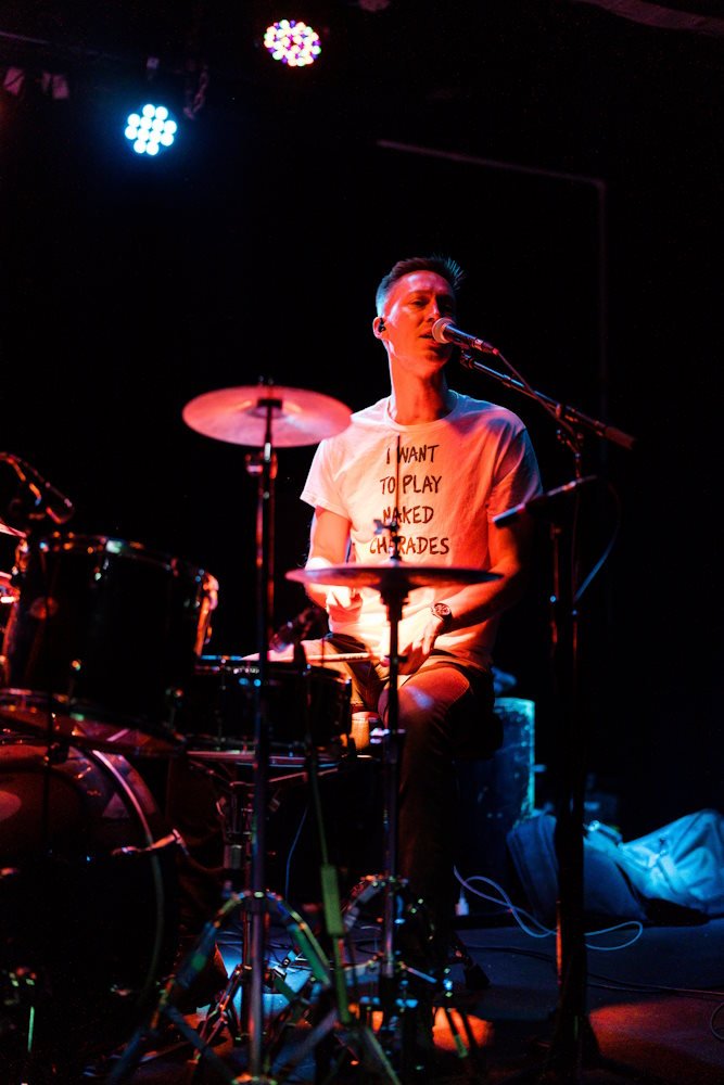 Person playing drums and singing on stage under colorful lights, wearing a T-shirt with text "I want to play naked charades."