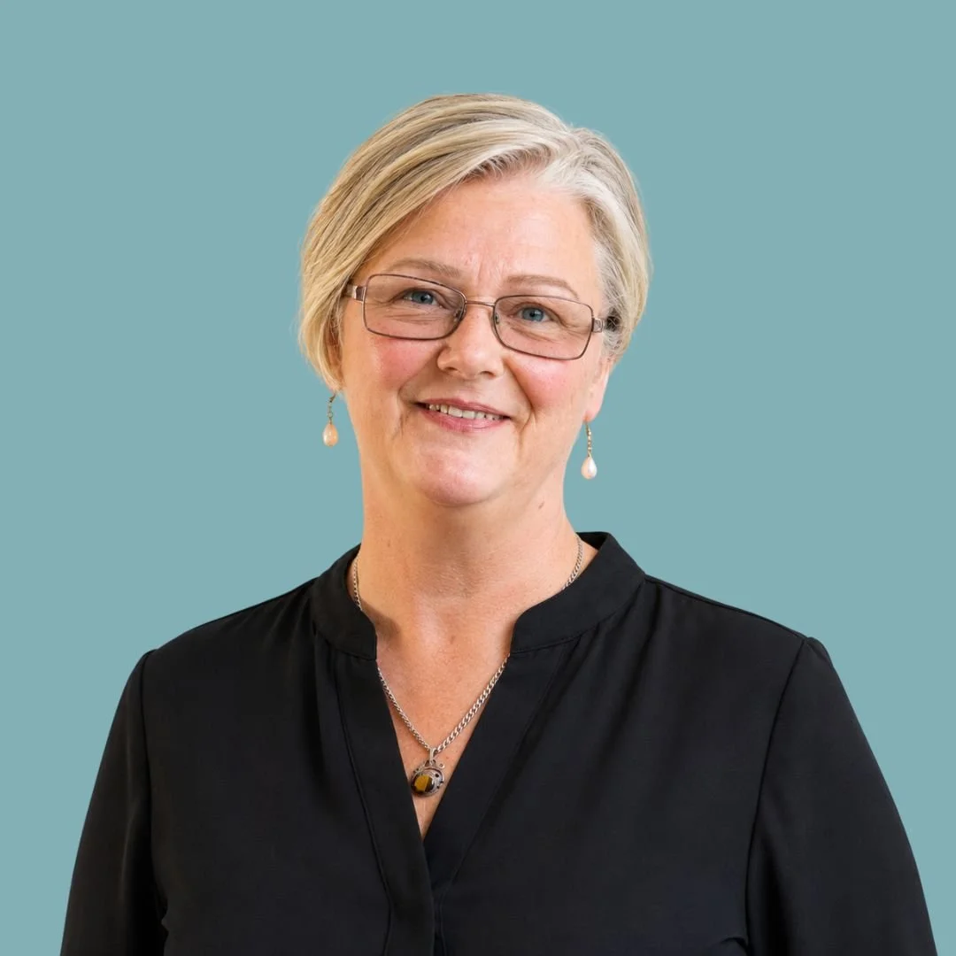 A smiling Jenny Mason, Complex Care Team Leader at UBH, with short blonde hair, wearing gold rimmed glasses, pearl earrings, and a bright pink and black patterned blouse, against a solid teal background.
