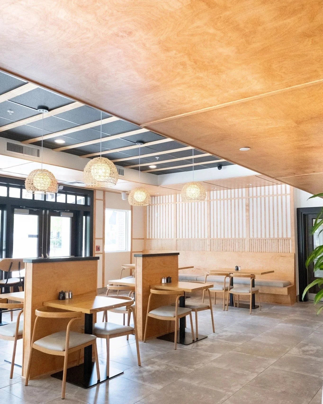 Interior of a modern restaurant with wooden furniture, hanging pendant lights, large windows, and a neutral color palette.