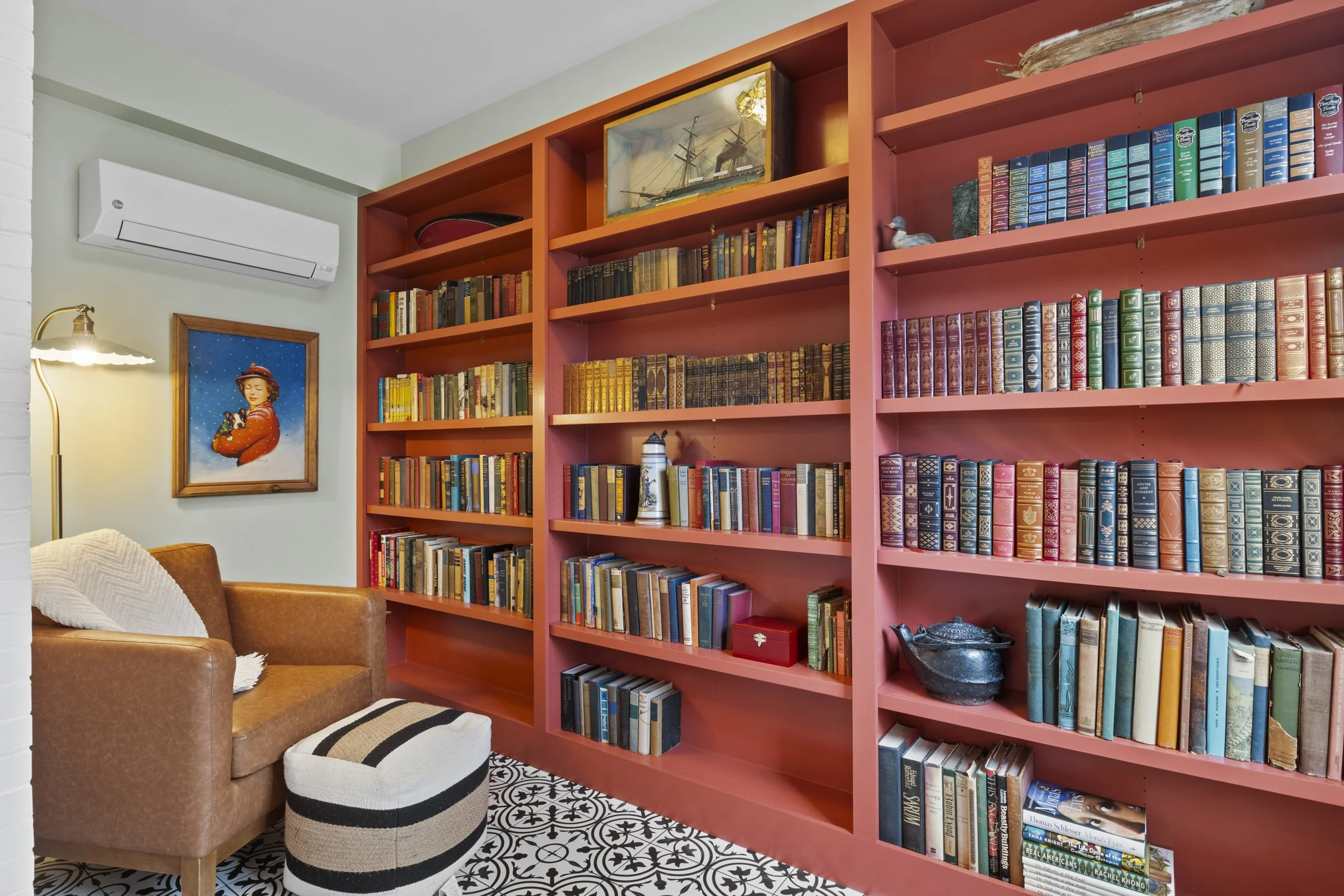 A living room corner with a brown leather armchair, a white and black striped pouf, a vintage floor lamp, a framed picture of a woman, and a tall pink bookshelf filled with books. The floor has black and white patterned tiles, and a white wall air co