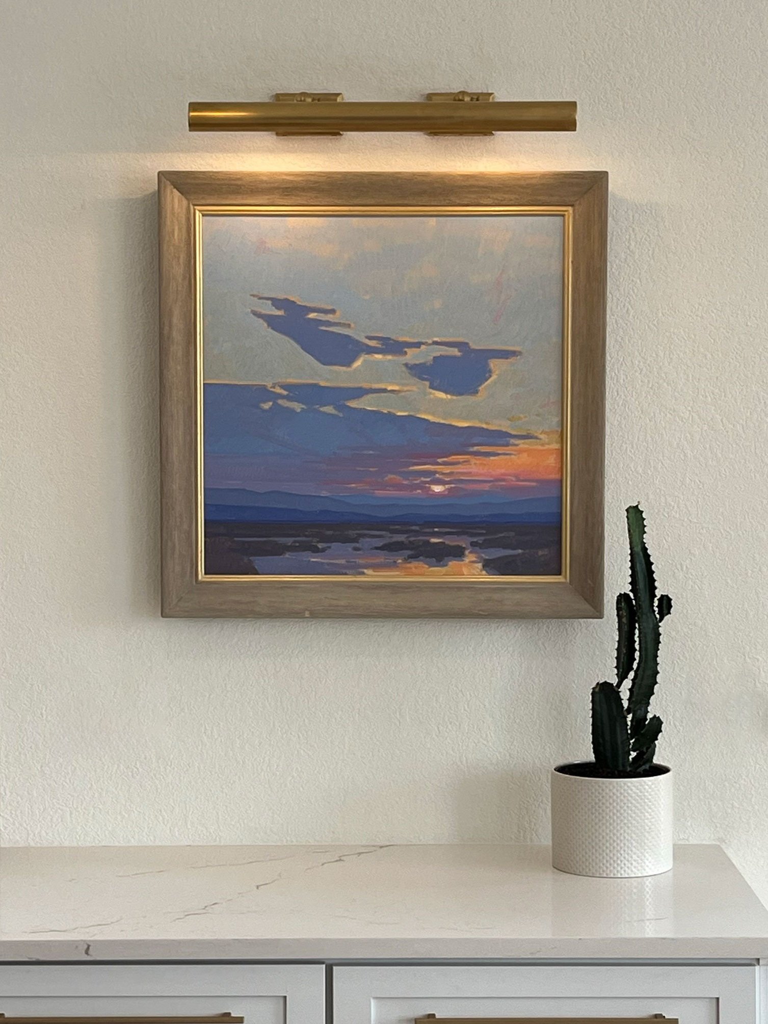 Painting hanging in kitchen with art light and cactus on the counter