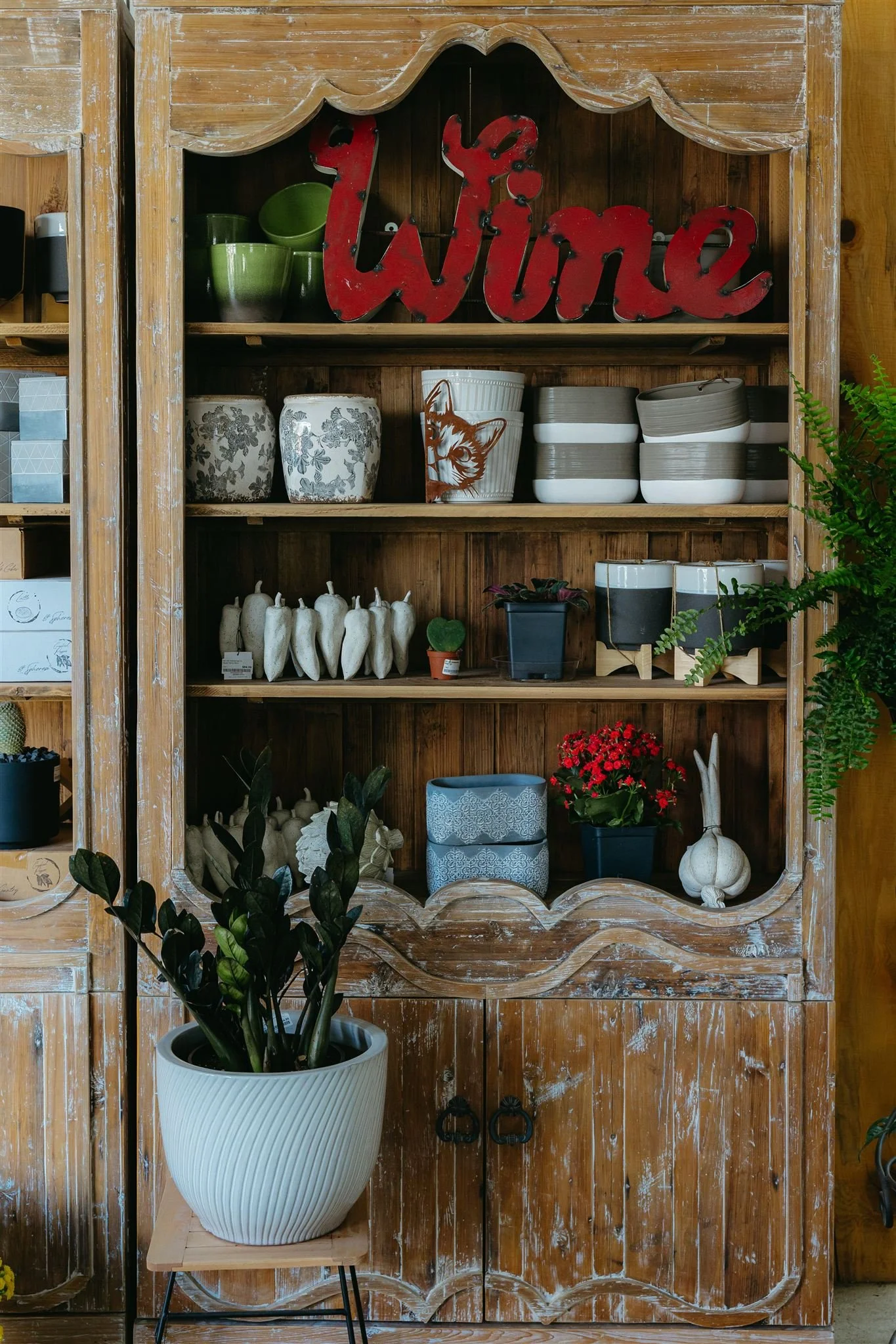 Decorative wooden cabinet with plants and pottery, including a large white potted plant in the foreground, and a red "Wine" sign on the top shelf.