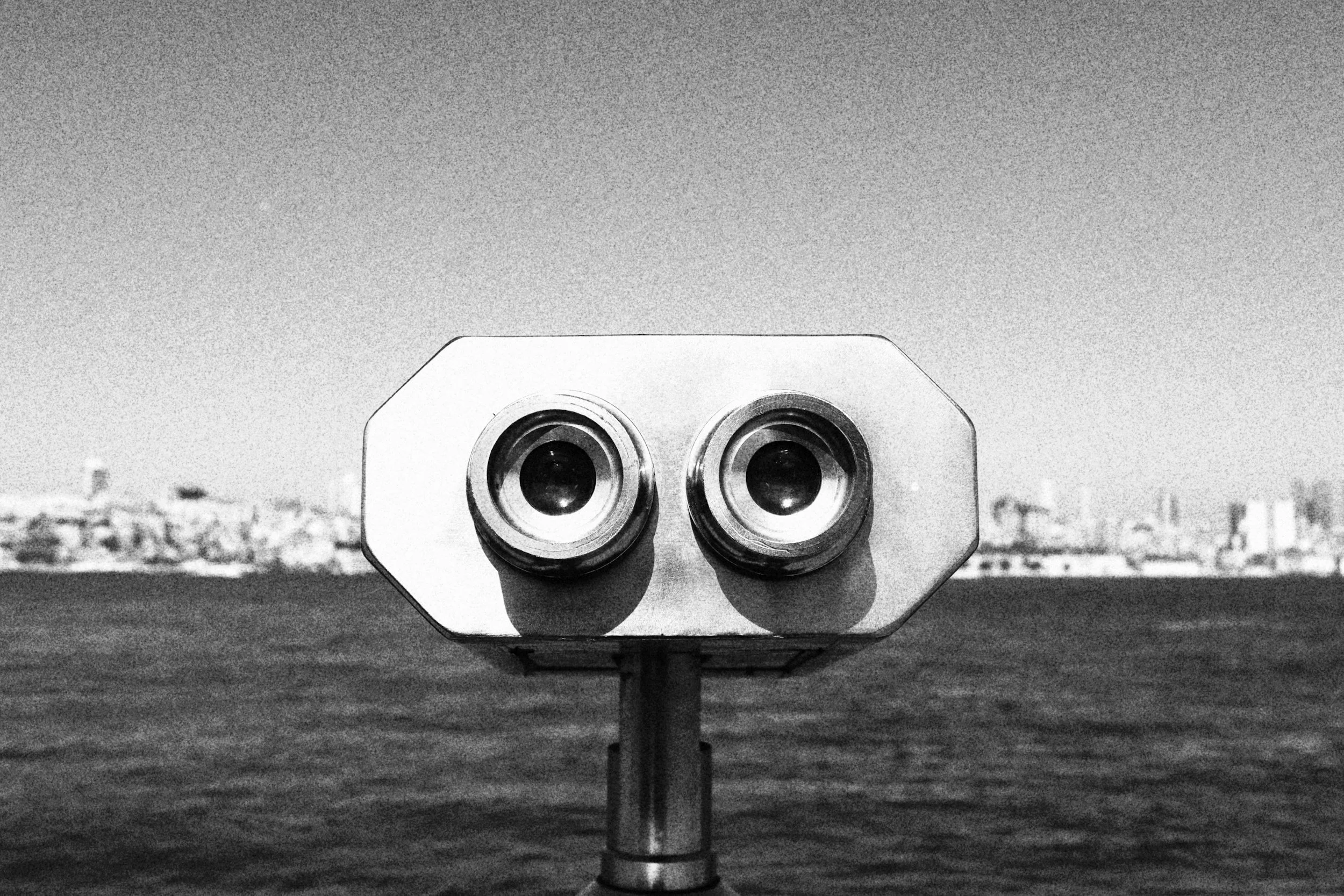 A black and white photo of a coin-operated binocular viewer overlooking a body of water with city buildings in the distance.