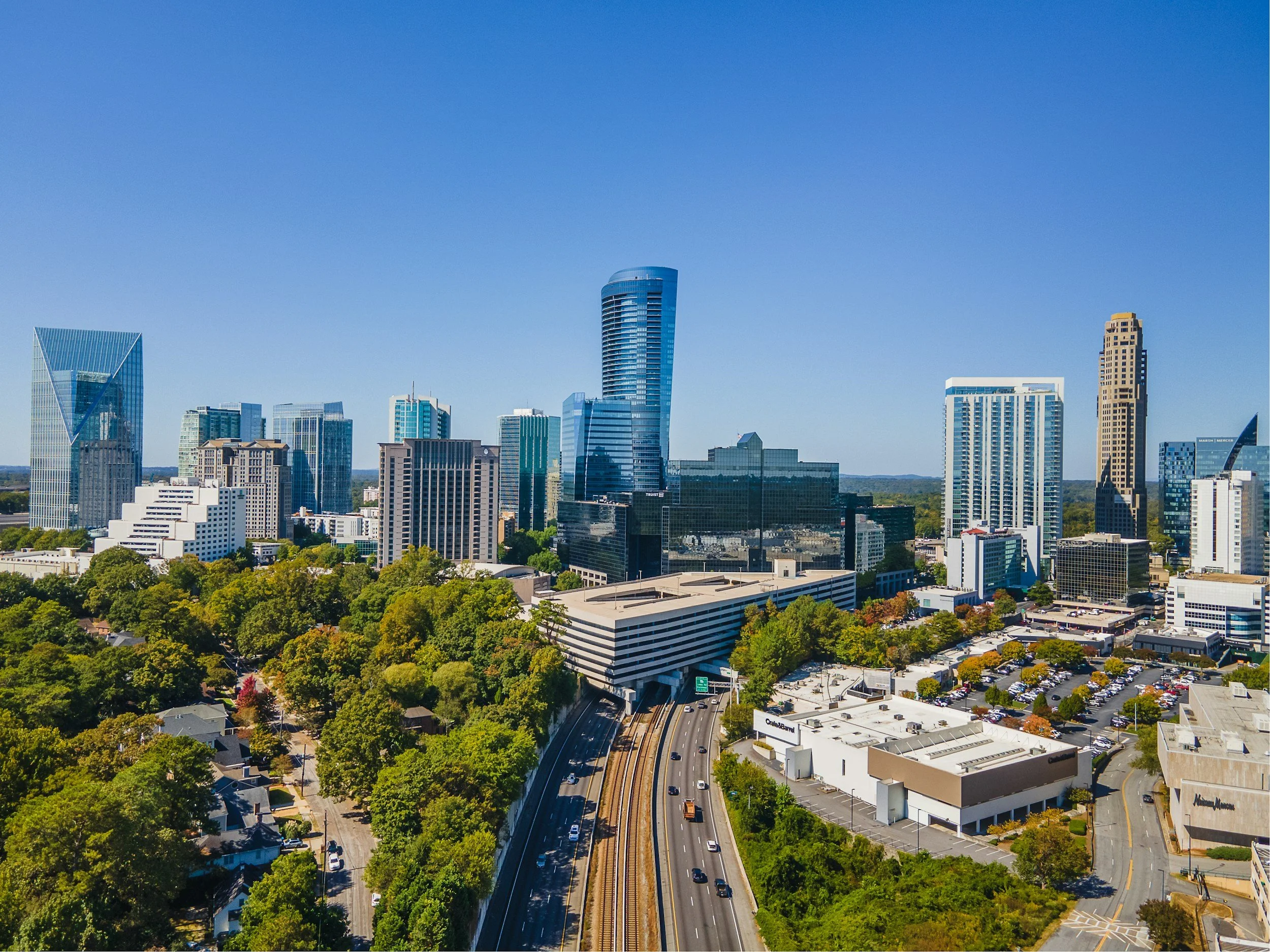 A city skyline featuring tall modern skyscrapers, with a clear blue sky overhead and a highway with cars and train tracks in the foreground, surrounded by green trees and low-rise buildings.