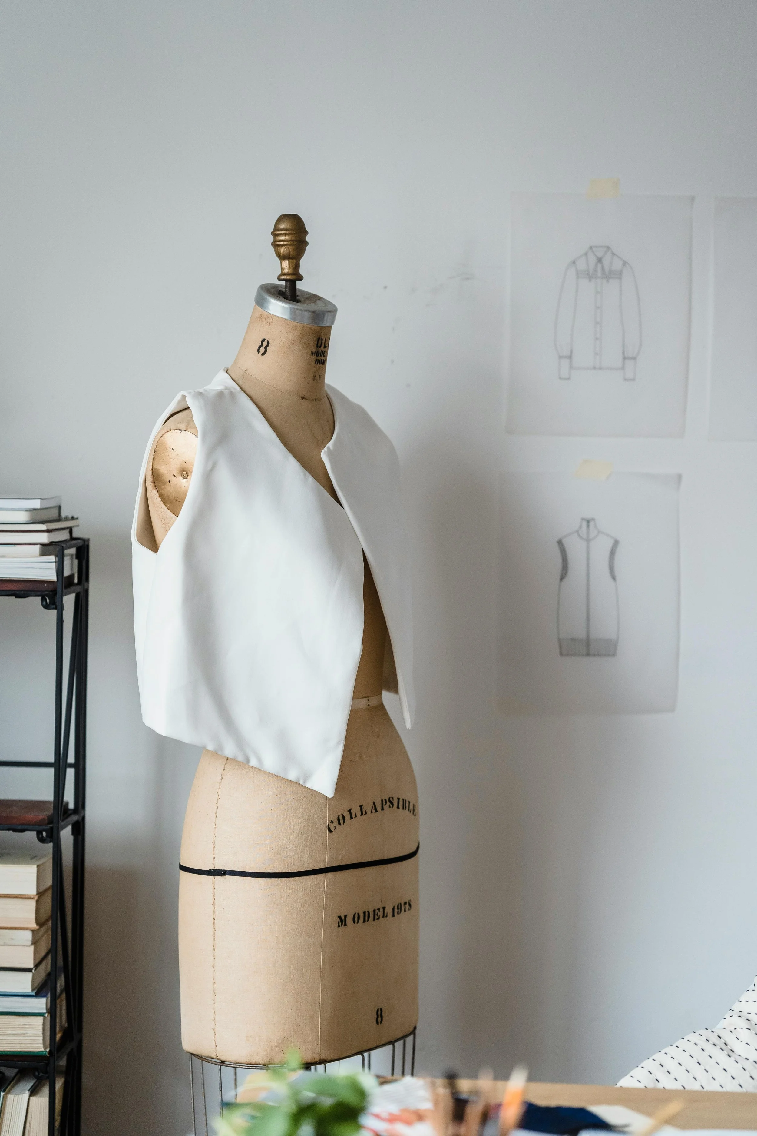 A dress form mannequin with a white sleeveless vest on it, set in a studio or design workspace with sketches of clothing on the wall.