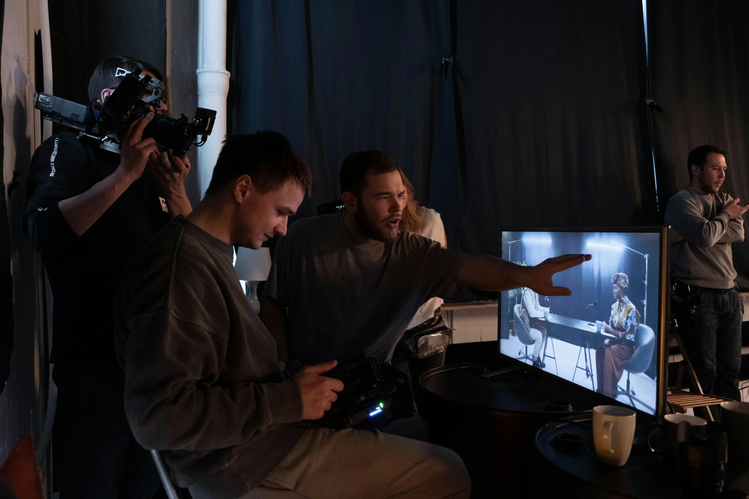 Filmmakers on set filming a scene with two women in a studio, one with orange hair and one with long dark hair, sitting at a table, seen on a monitor.