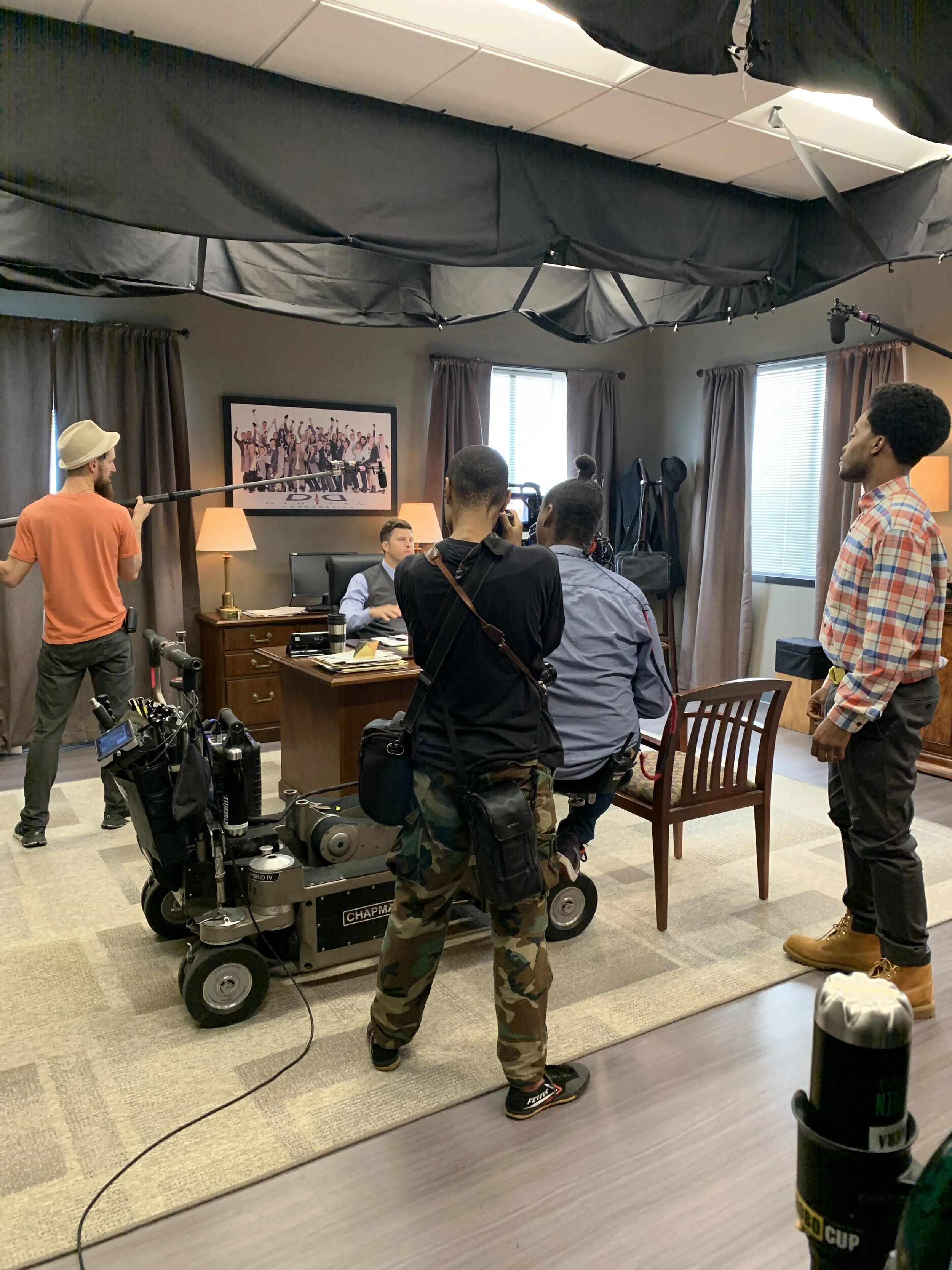 Filming crew setting up a scene with actors in an office, with camera equipment, crew members, and a director or producer at the desk.