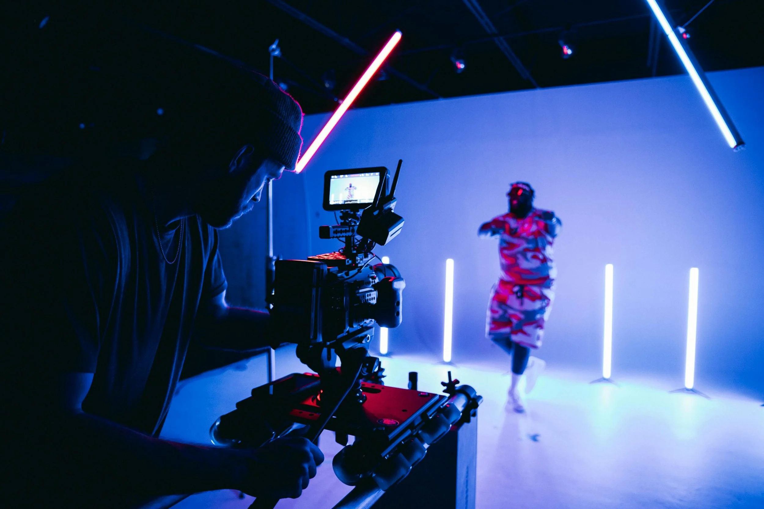Filmmaker operating camera in a studio with neon blue and pink lighting, a person in trendy clothing posing in the background.