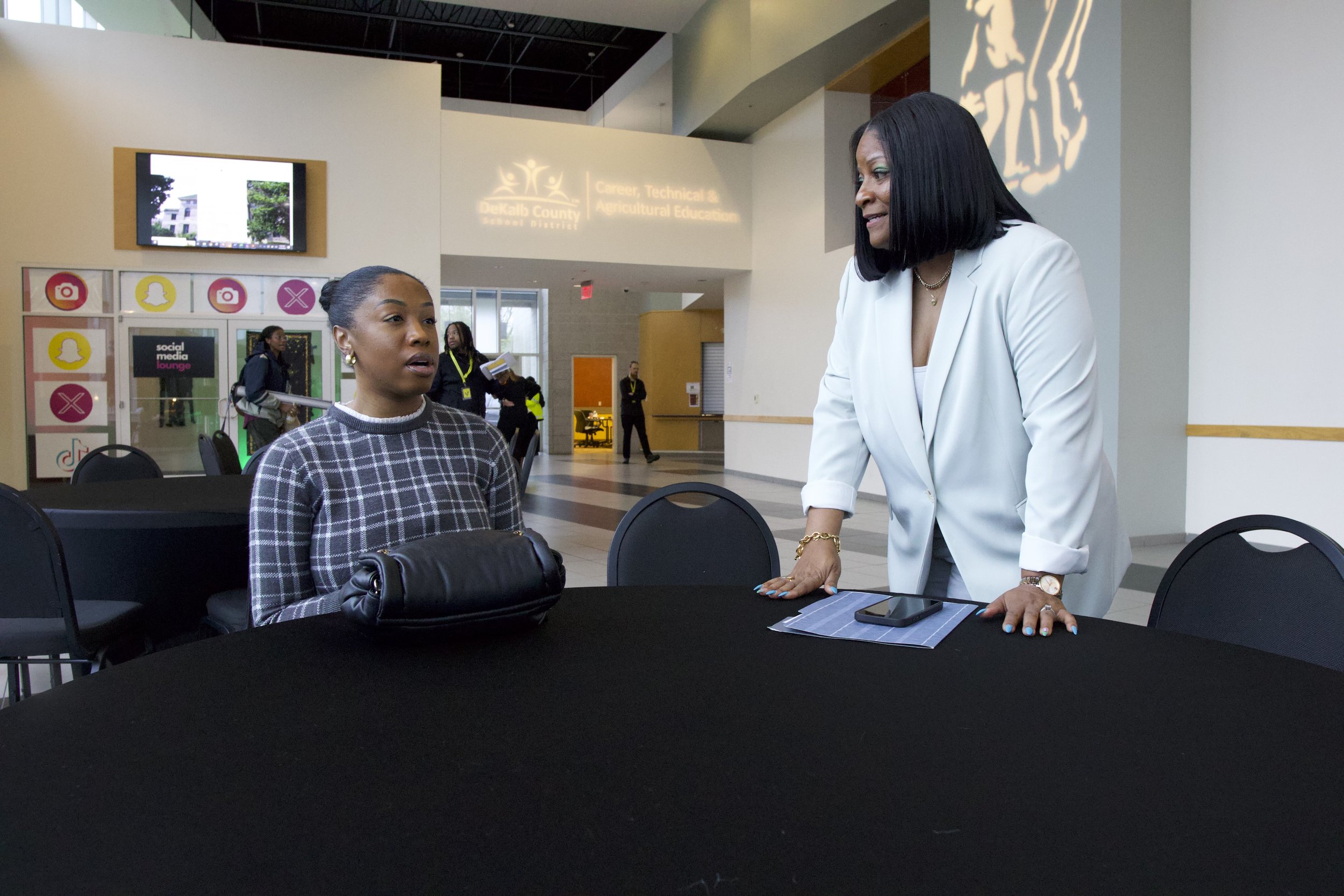 Two women having a discussion at a table in an indoor public space, with media icons in the background.