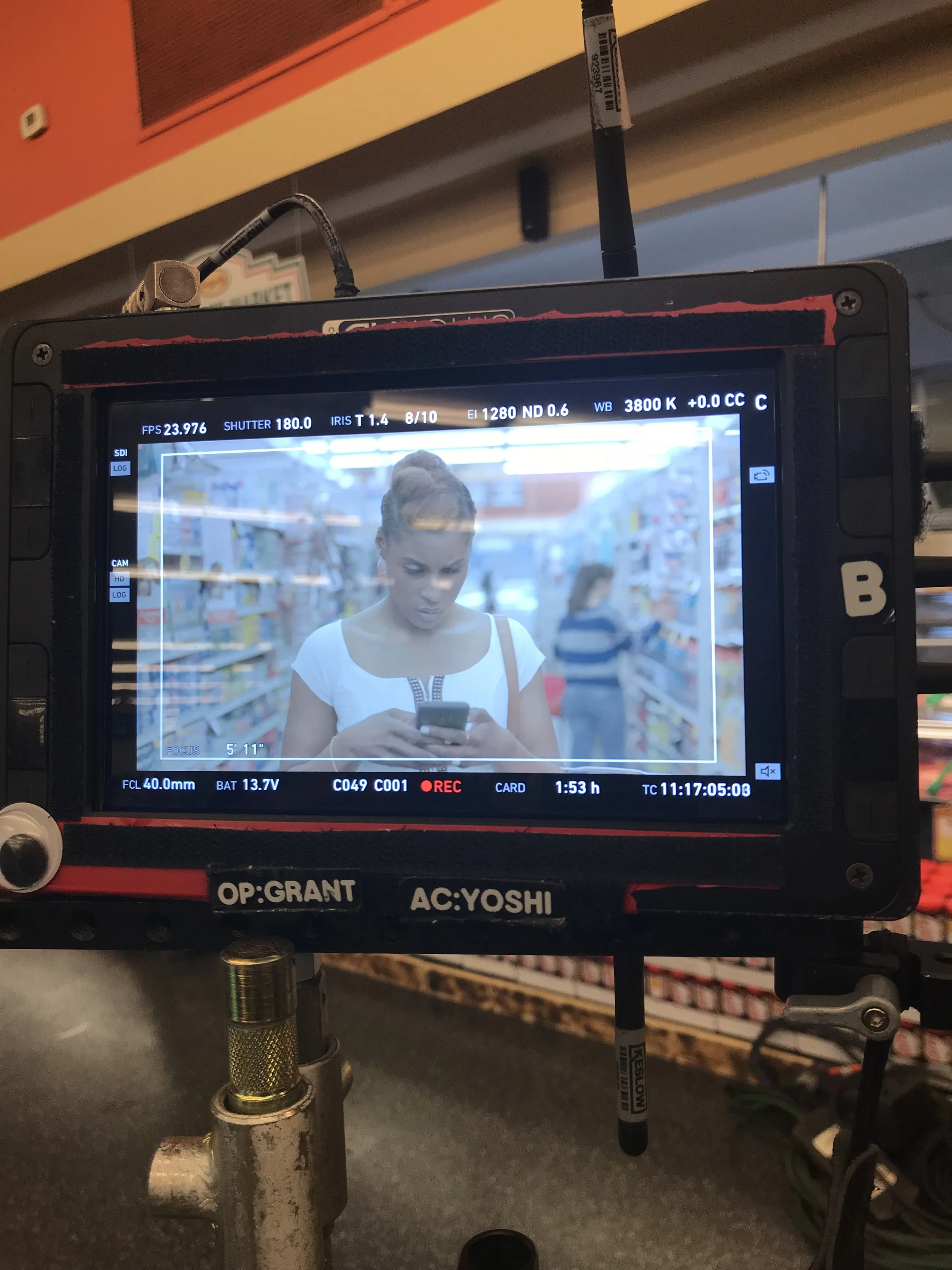 A woman in a grocery store appears on a camera monitor, looking at her phone with shelves of products behind her.