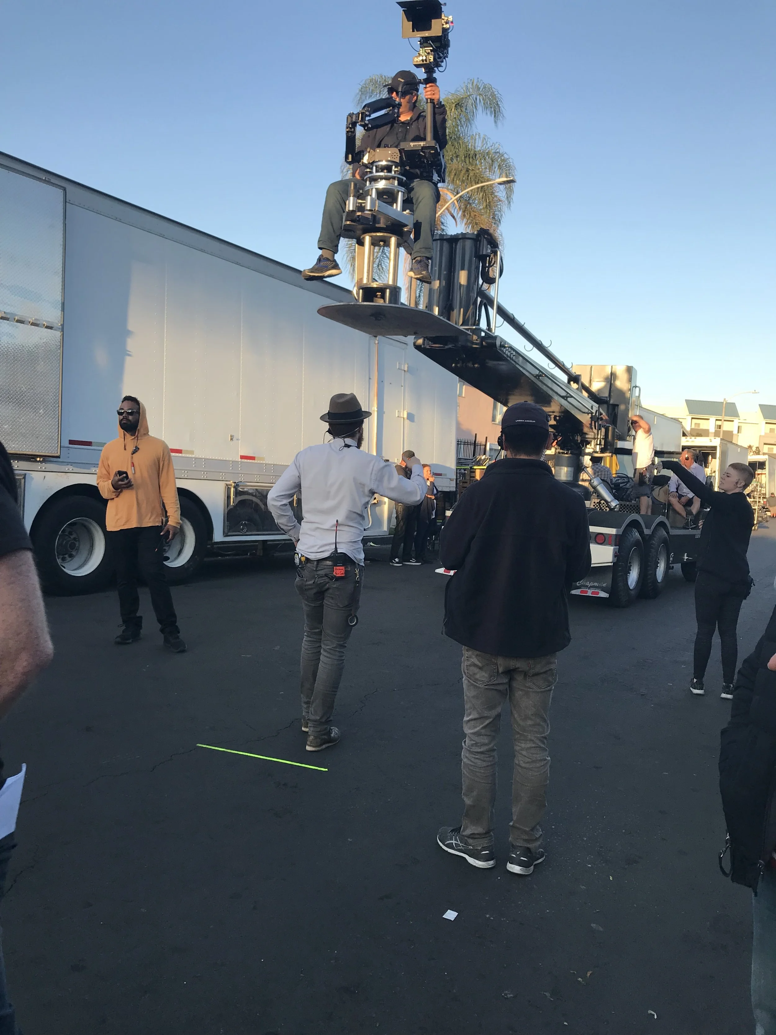 A film crew working outdoors with a camera crane, people gathered around, some observing and others working, on a street with buildings and a clear sky.