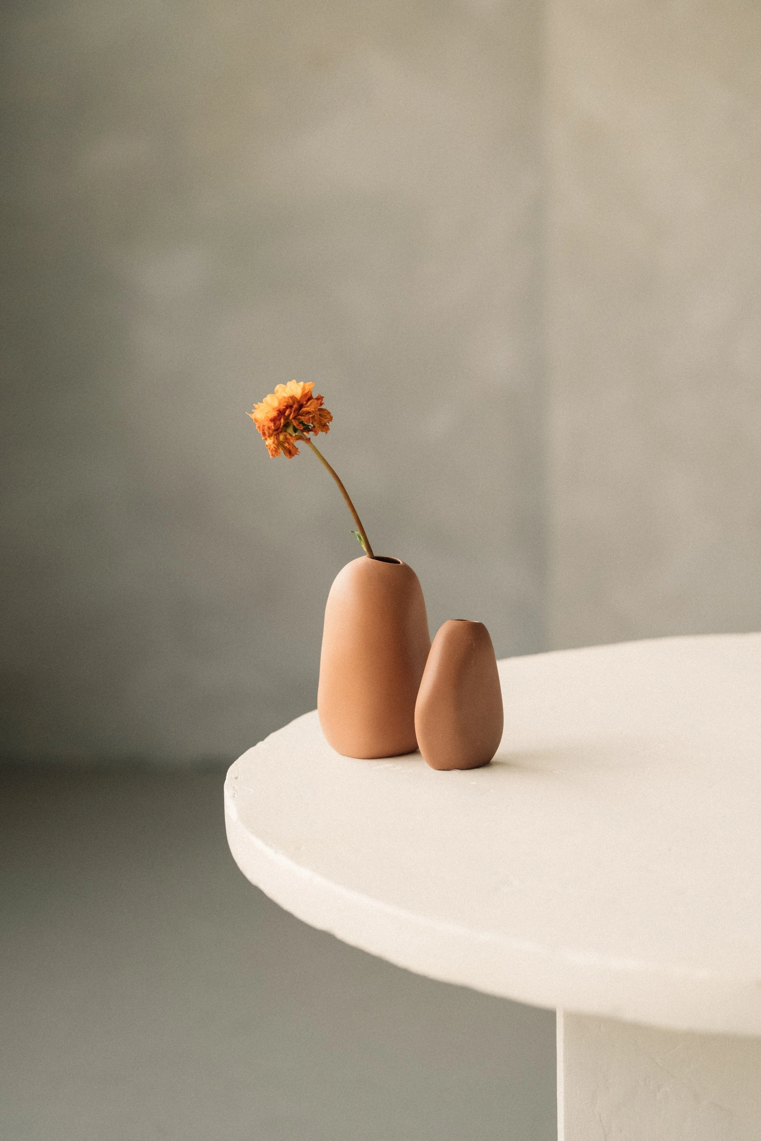 Two brown vases, one taller with an orange flower, sit on a white round table against a neutral background.