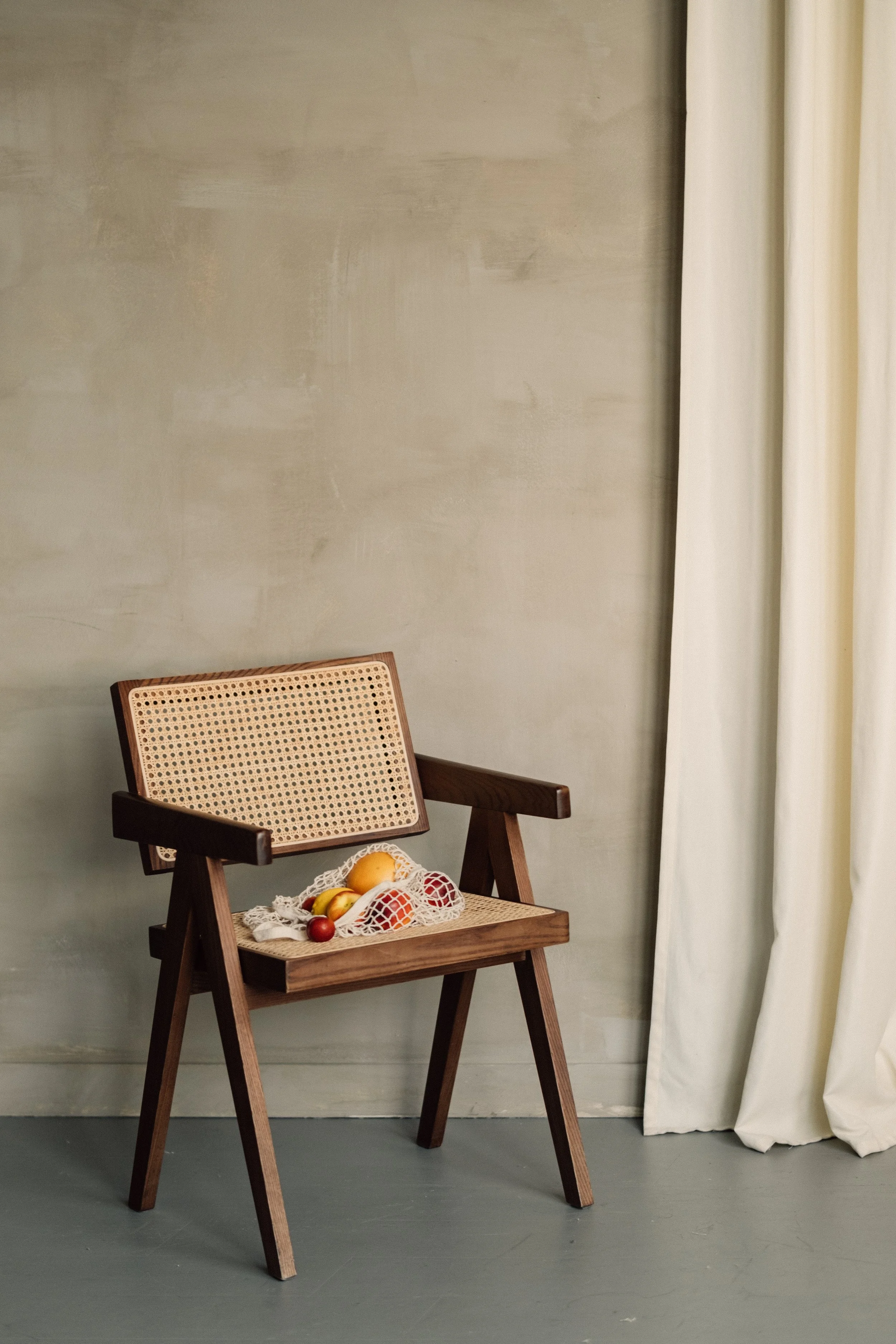 Wooden chair with a cane backrest and seat, with a small mesh bag of assorted fruits resting on it, positioned in a room with a plain wall and white curtains.