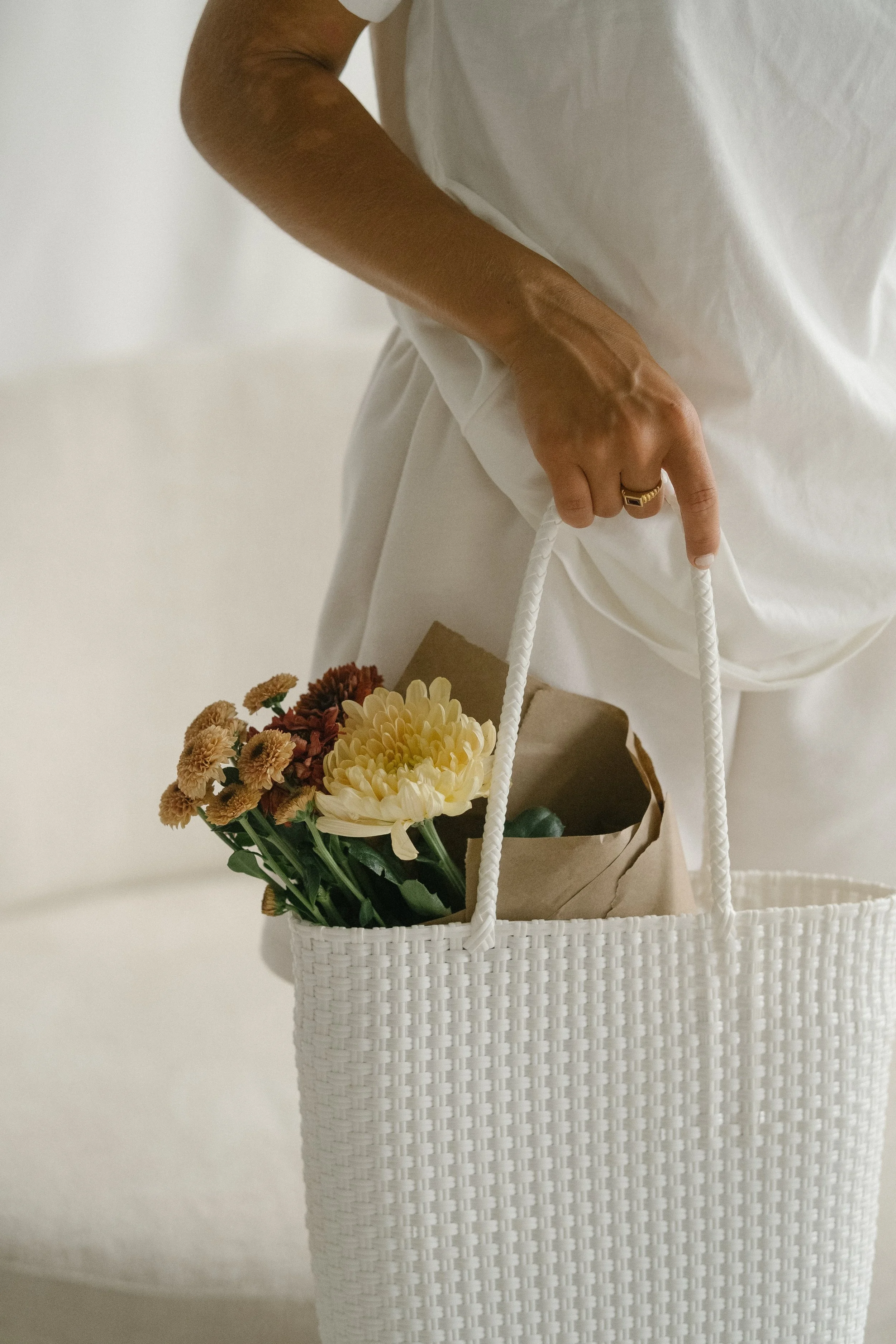 Person holding a white woven bag with flowers inside, wearing a white outfit, in a neutral background setting.
