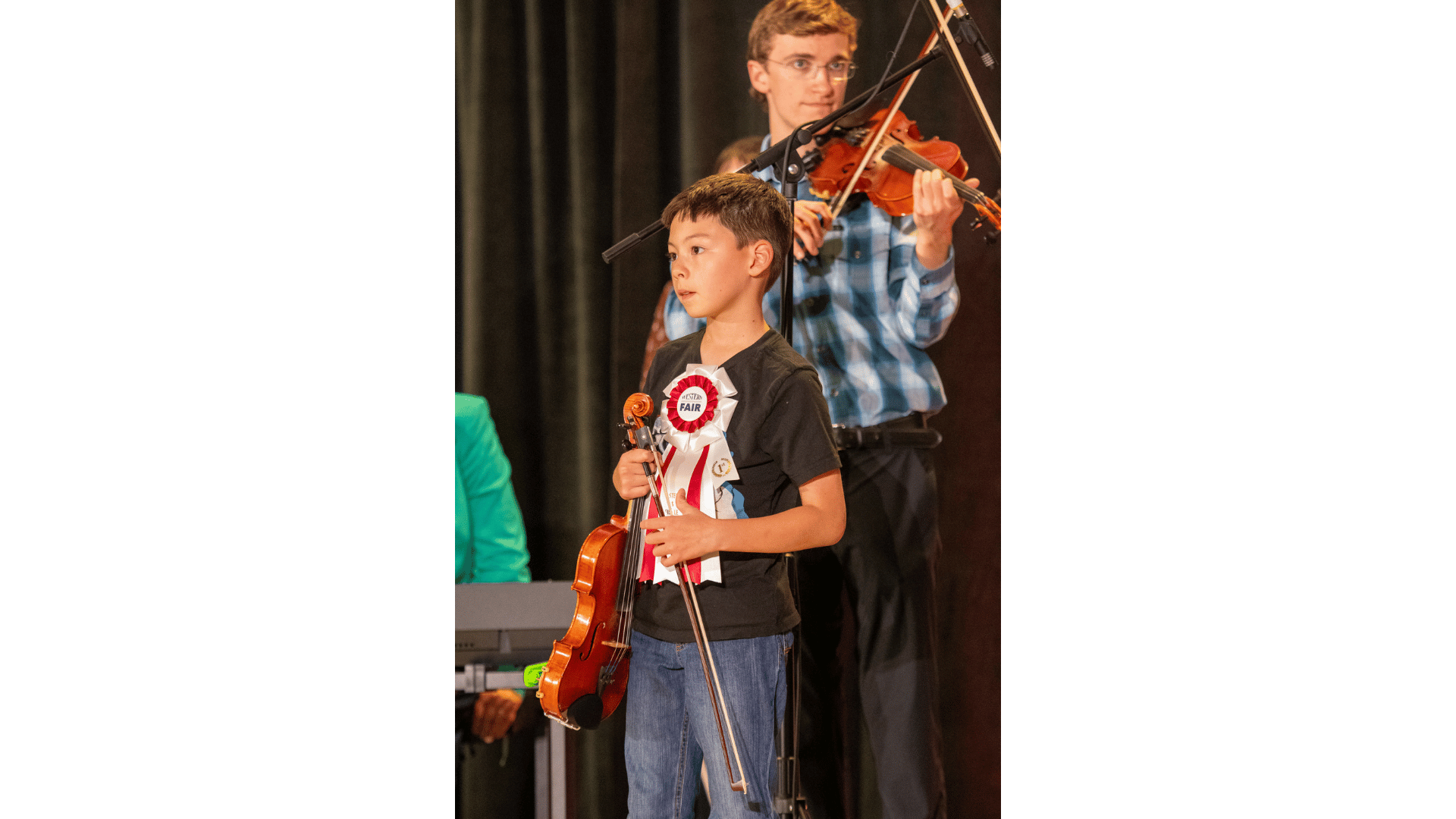A young boy holding a violin and a bow, wearing a ribbon that says 'Fair,' standing on stage during a musical award ceremony. Behind him, a man is playing a violin, seen wearing glasses and a checkered shirt.