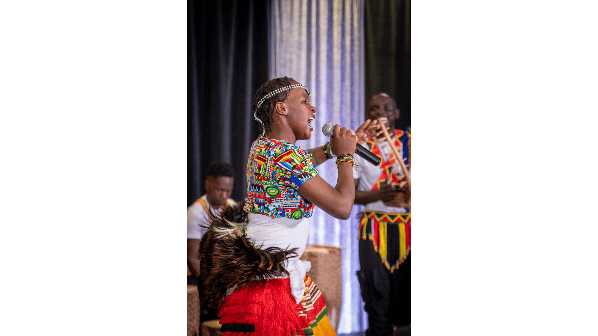 Young woman singing into a microphone during a cultural performance, wearing traditional colorful attire and headband, with men in similar attire playing instruments in the background.