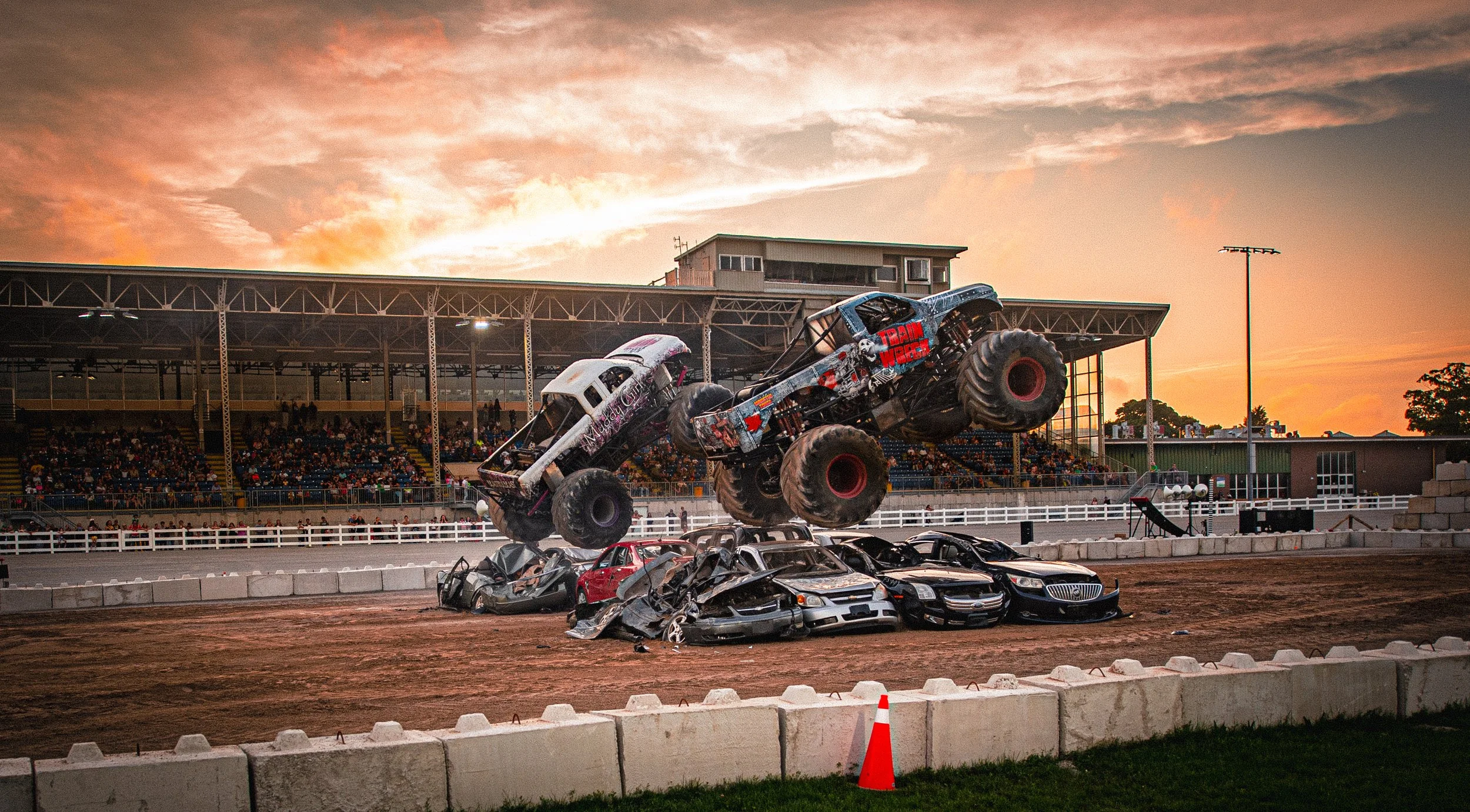 A monster truck with large tires and a customized paint job jumping over crushed cars on a dirt track at a fairground during sunset, with spectators in the stands.