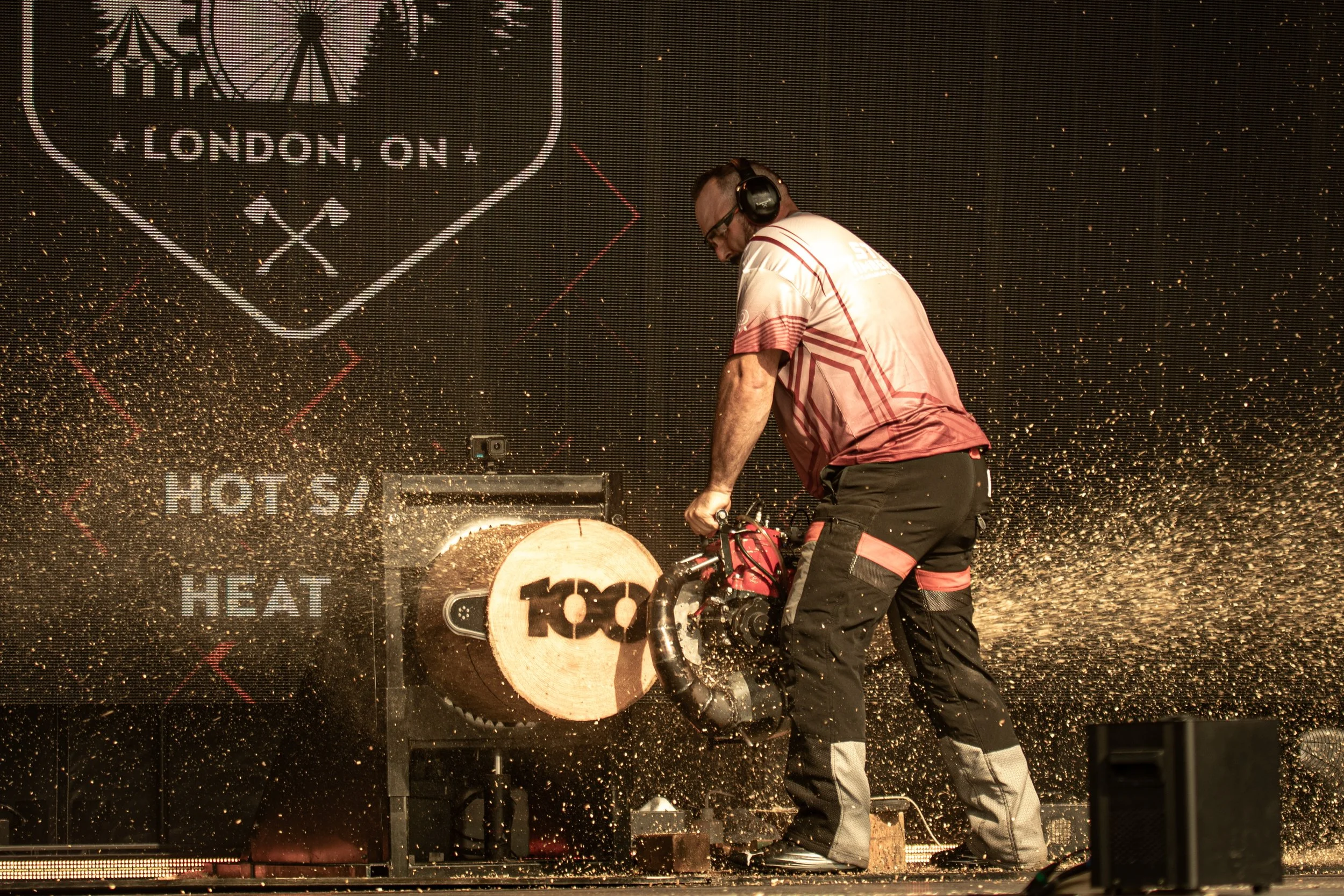 A person wearing headphones and safety glasses operating a wood-cutting saw on a piece of wood, with sparks flying around in a workshop or stage setting.
