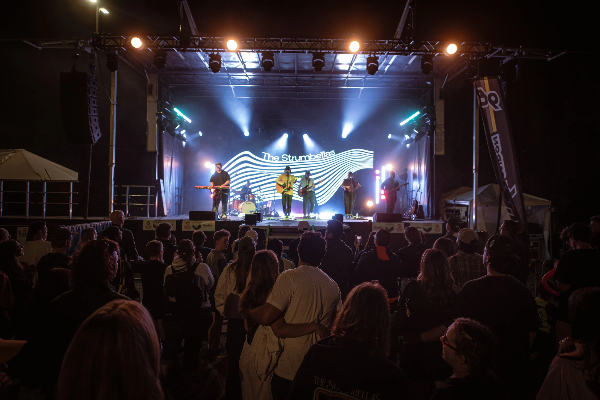 A live music concert with a band performing on stage under stage lights, with an audience watching at night.
