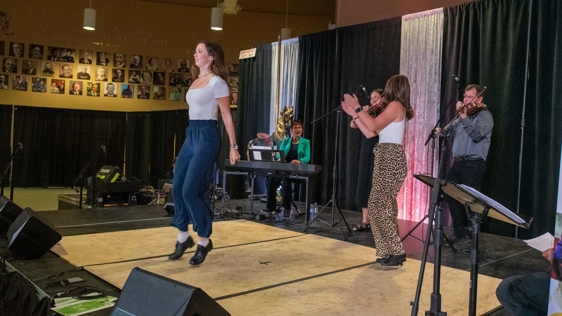 Performer dancing on stage with musicians and a woman in a green blazer and a woman in leopard print pants clapping nearby, in an indoor venue with black curtains and photos on the wall.