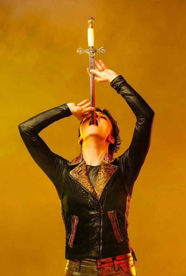 Person in black leather jacket balancing a sword vertically with the tip in their mouth against a yellow background.
