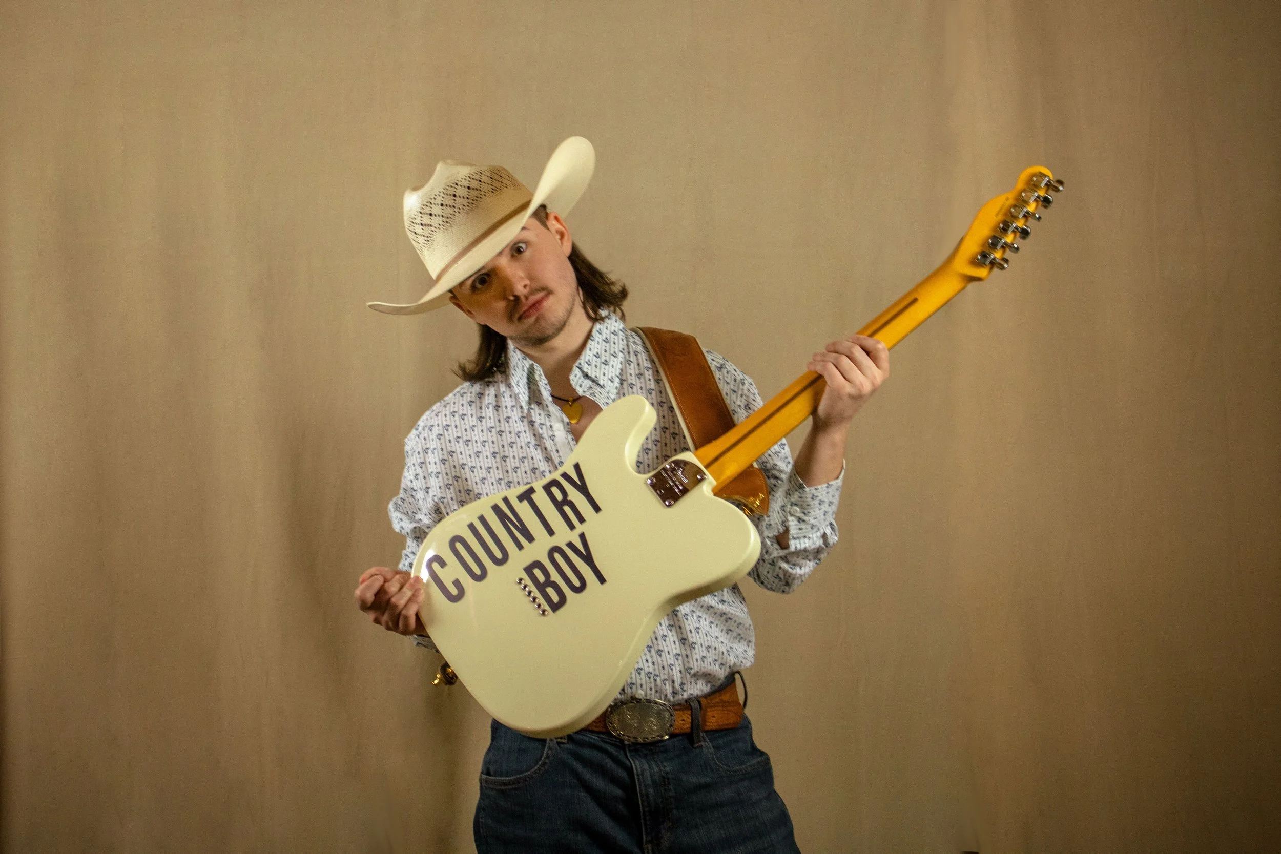 A man wearing a white cowboy hat, patterned shirt, and jeans holding a cream-colored electric guitar that has a sign reading 'COUNTRY BOY' on it. He is standing against a plain beige wall.