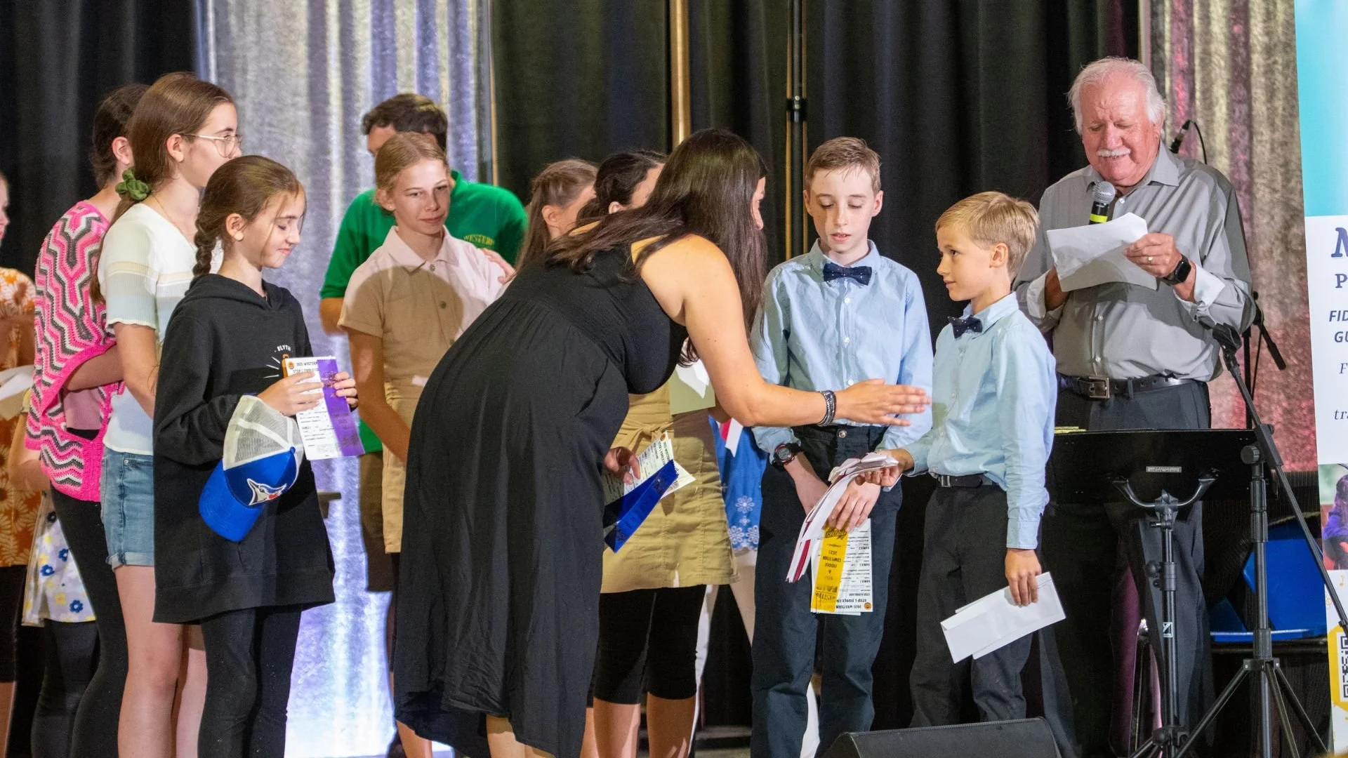 A group of children and adults on stage receiving awards or recognition. An older man is reading from a paper, while a woman speaks to two boys in front of her. The children are holding certificates and gifts, and the event appears formal with an audience.