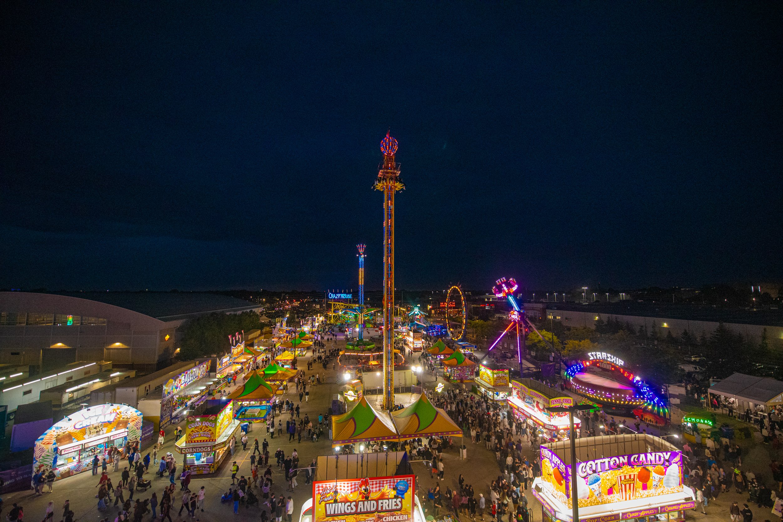 A nighttime view of a colorful carnival with rides, food stalls, and bright neon signs, filled with visitors walking around.
