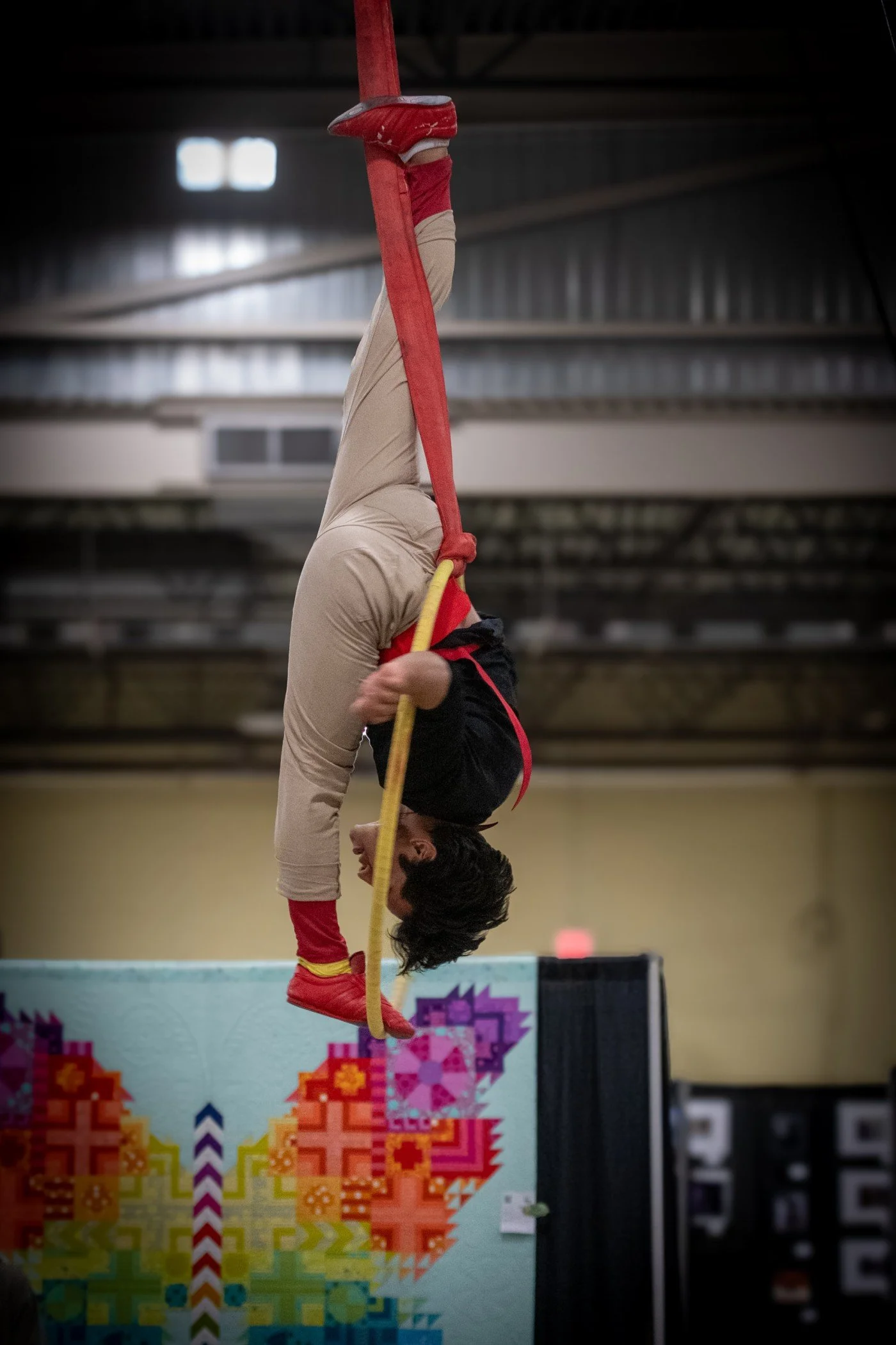 A person with long dark hair performing a handstand on a slackline inside a gymnasium. The person is wearing beige pants, a black shirt, and red shoes. The background includes pixelated rainbow-colored artwork and industrial ceiling features.
