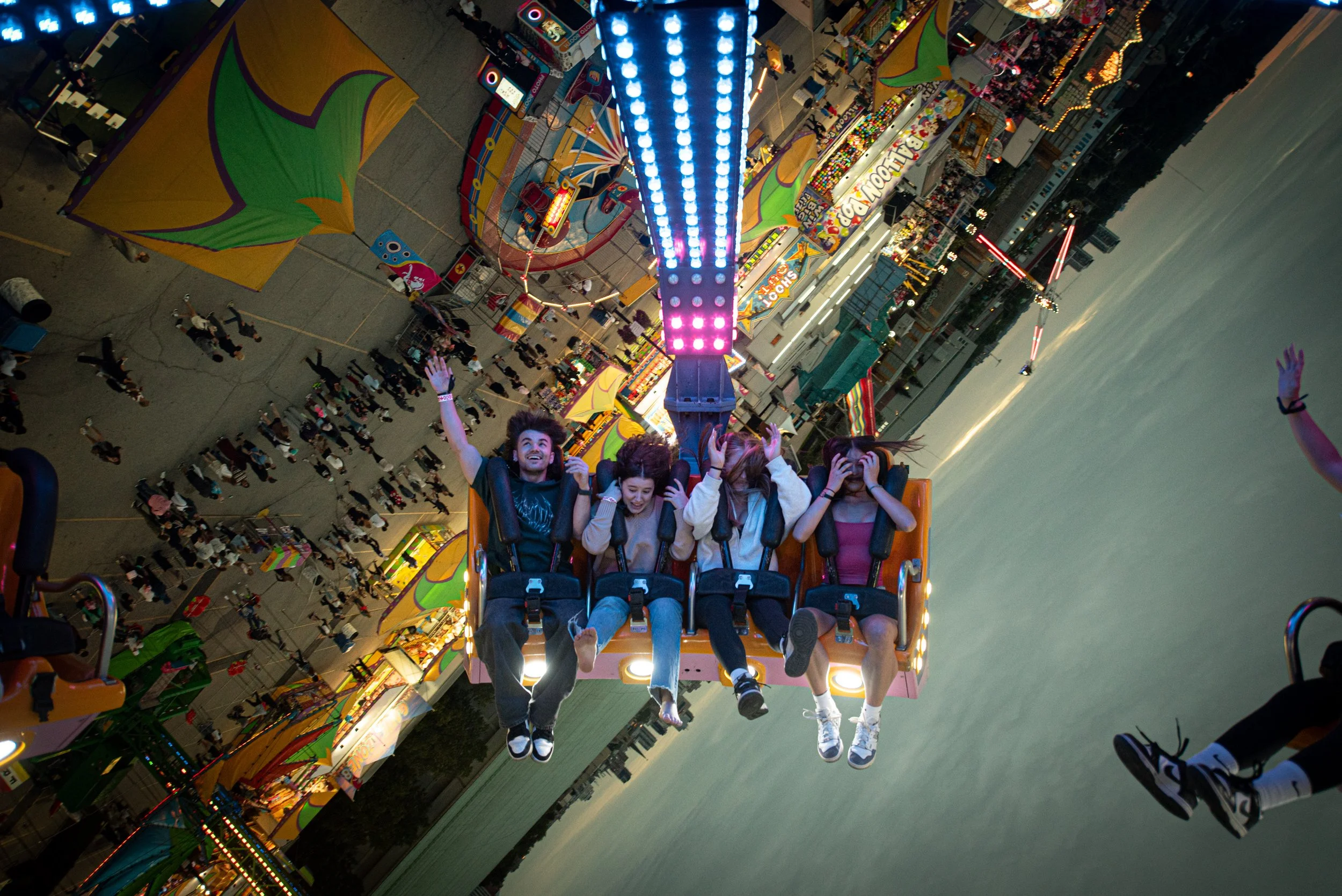 People riding a colorful amusement park ride, possibly a swing or drop tower, during the evening with amusement booths, flags, and city buildings in the background.