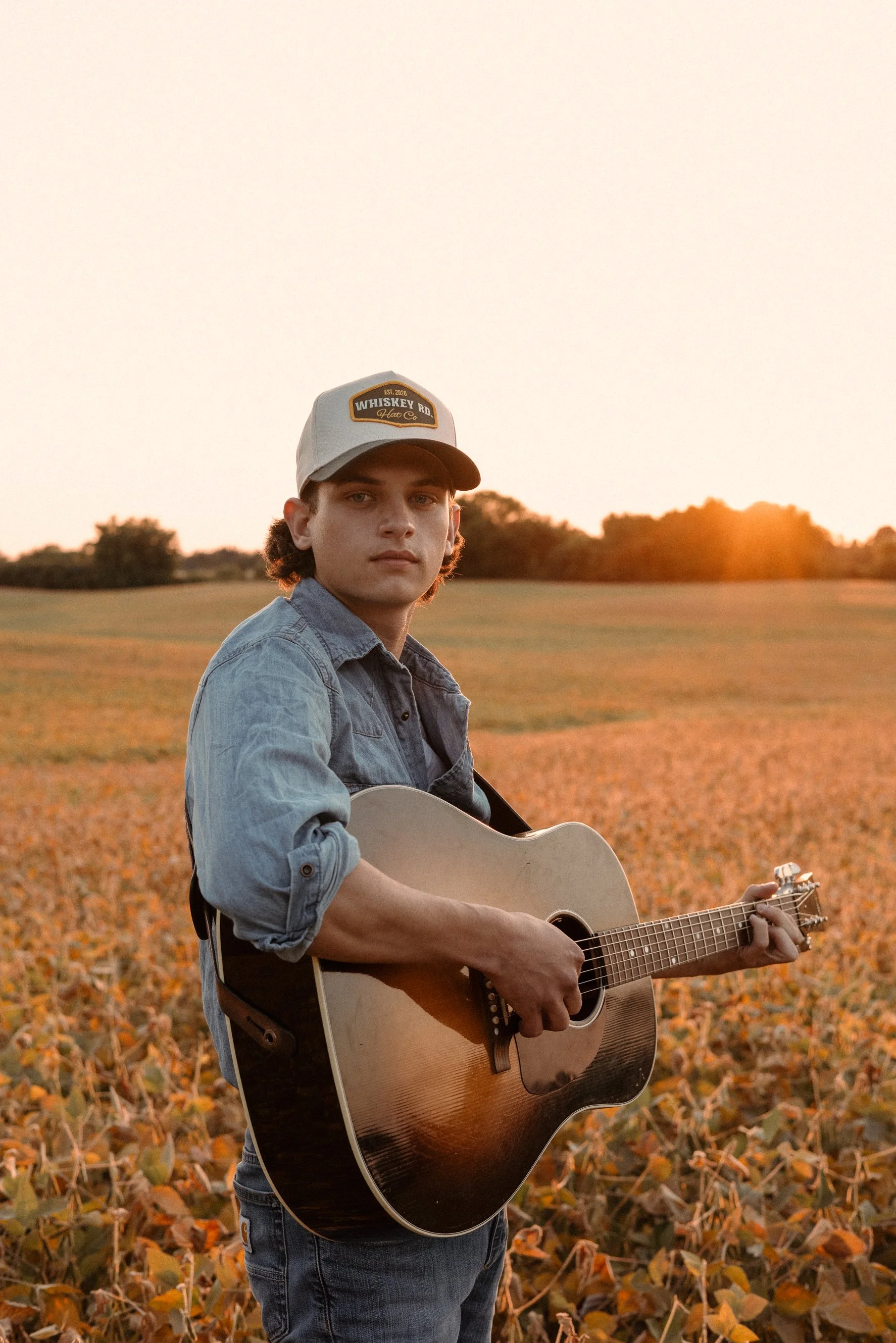 A young man in a denim jacket and cap playing an acoustic guitar outdoors during sunset, standing in a field with autumn leaves.