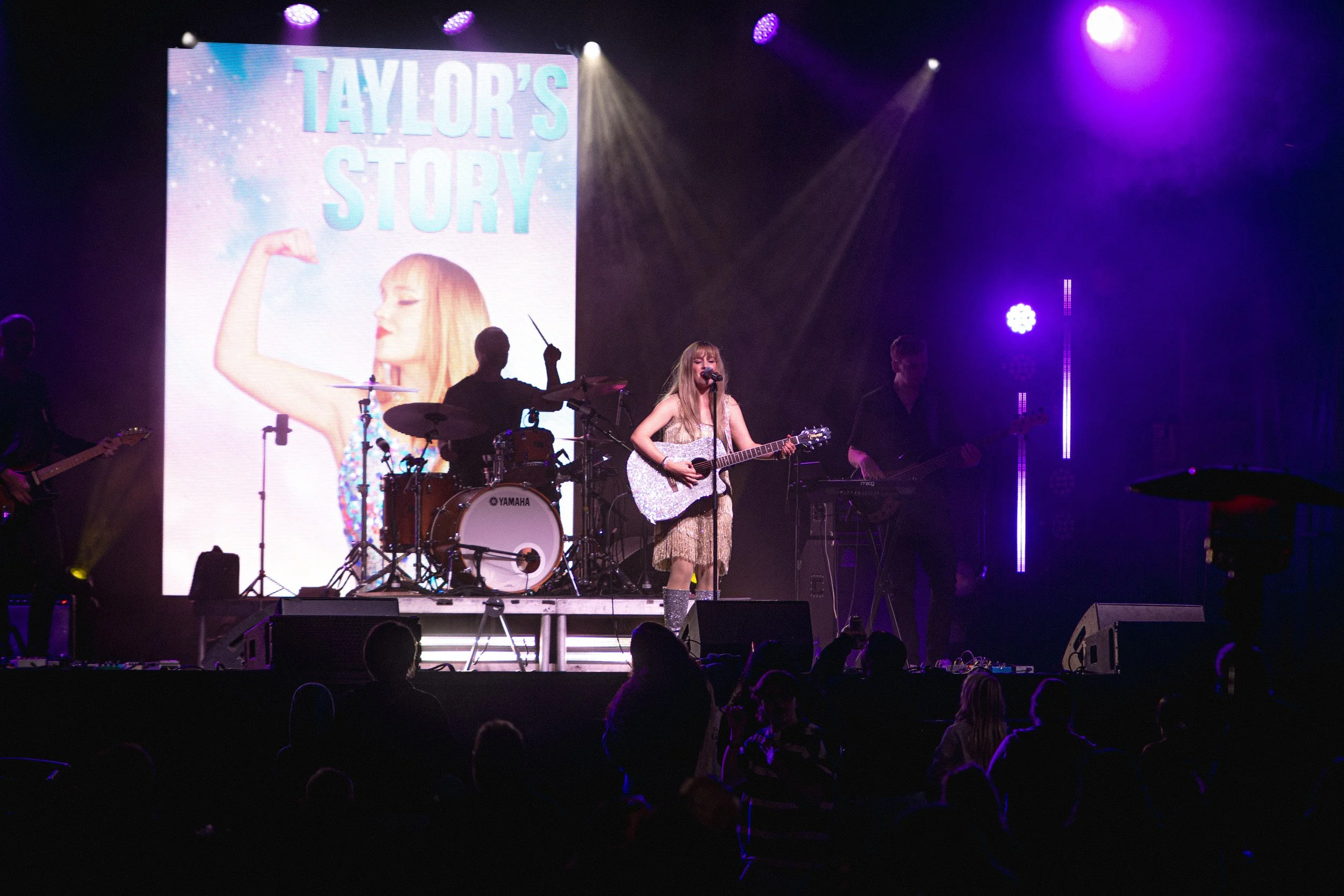 A musical performance on stage with a singer playing guitar, band members, and a large screen displaying 'TAYLOR'S STORY' behind them, lit with colorful stage lights.