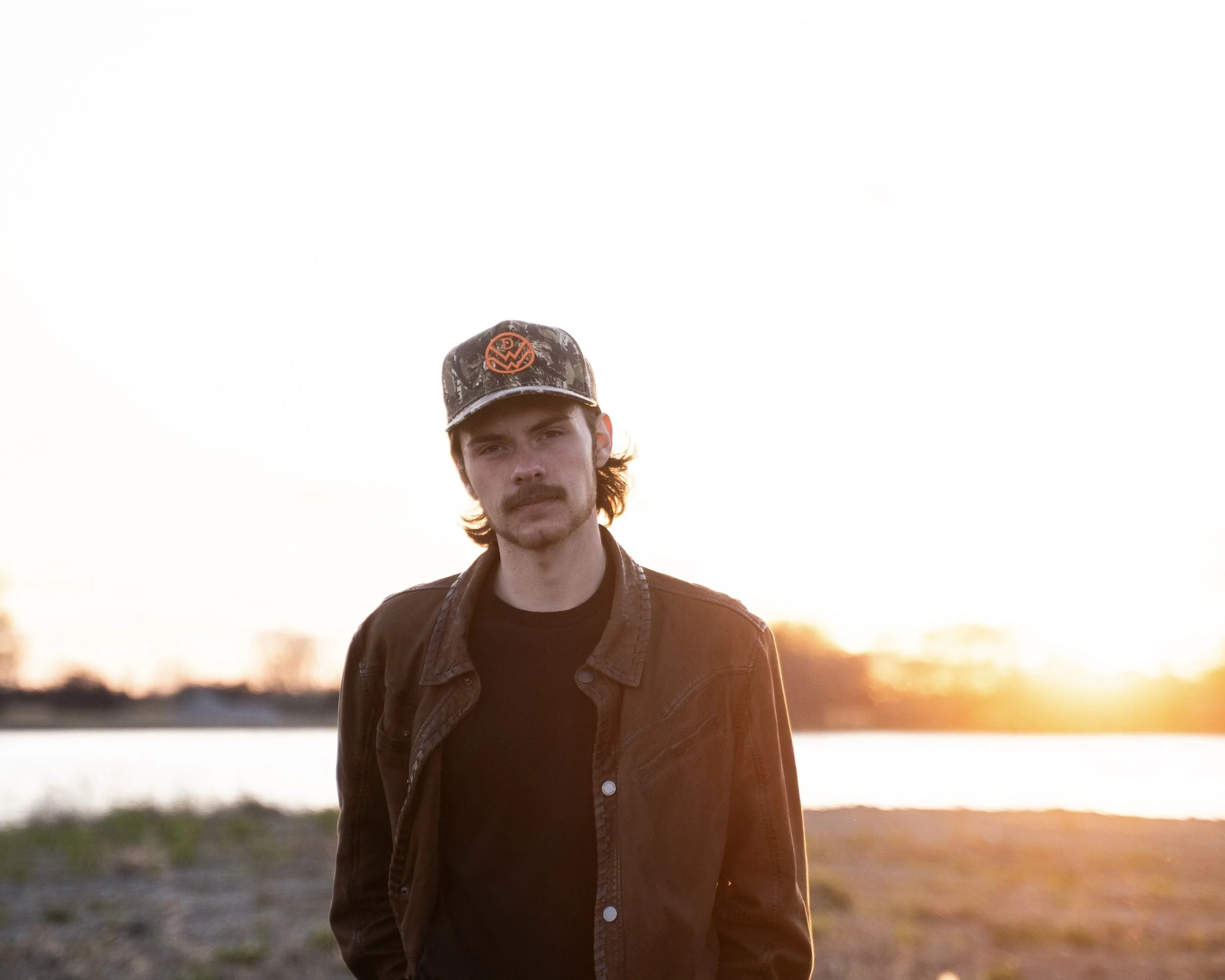 Young man with dark hair, mustache, and beard wearing a black jacket and camo hat standing outdoors during sunset.