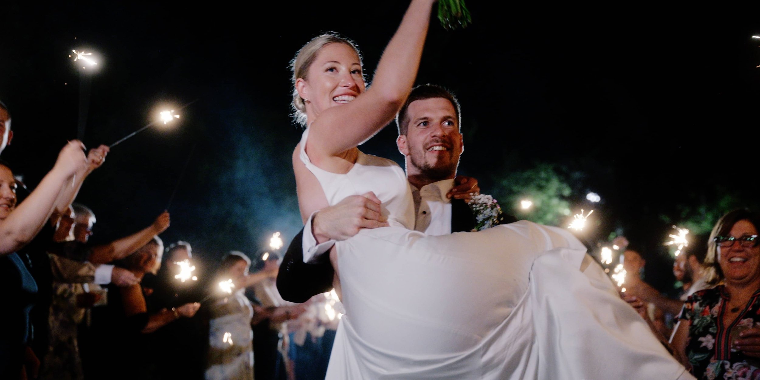 Couple celebrating at a wedding, the bride being lifted by the groom, surrounded by guests holding sparklers at night.
