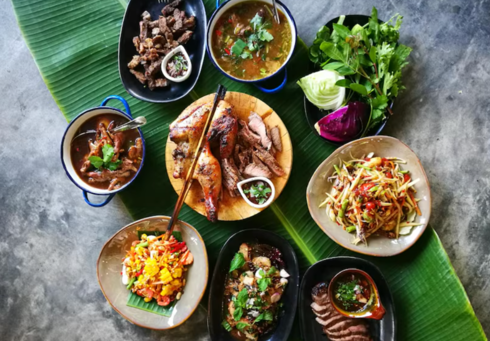 Assorted Thai dishes on a large green banana leaf, including grilled meats, papaya salad, and fresh herbs, served in bowls and plates.