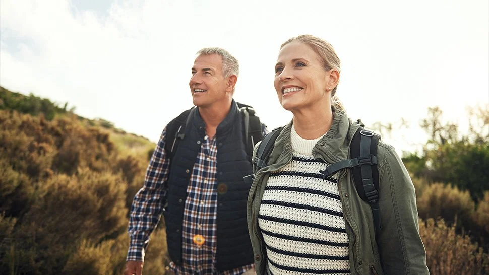 A smiling middle-aged man and woman hiking outdoors with backpacks, surrounded by nature and a partly cloudy sky.