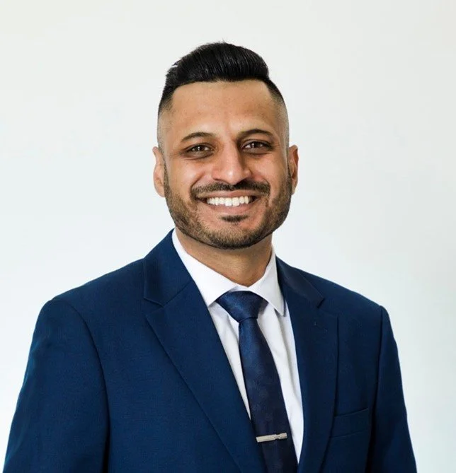 A man wearing a dark blue suit, white shirt, and striped tie, smiling and looking at the camera against a white background.