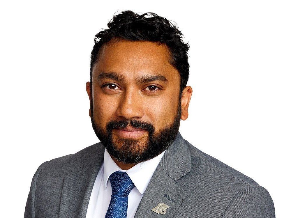 Professional portrait of a man with dark curly hair, beard, wearing a gray suit, white shirt, blue tie, and a lapel pin, against a white background.