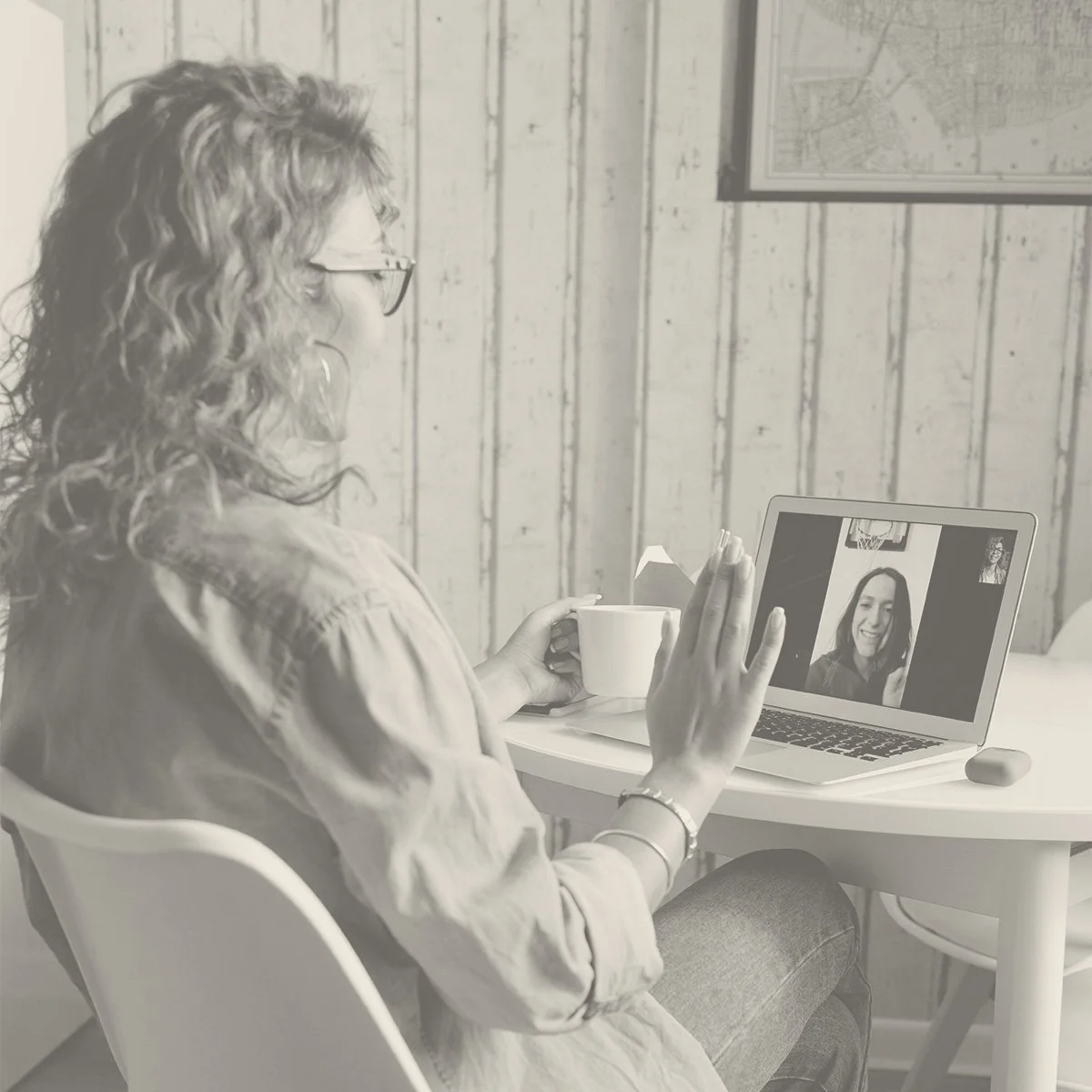 Woman with glasses and curly hair having a video call on a laptop, sitting at a white table with a coffee mug, in a rustic wood-paneled room.