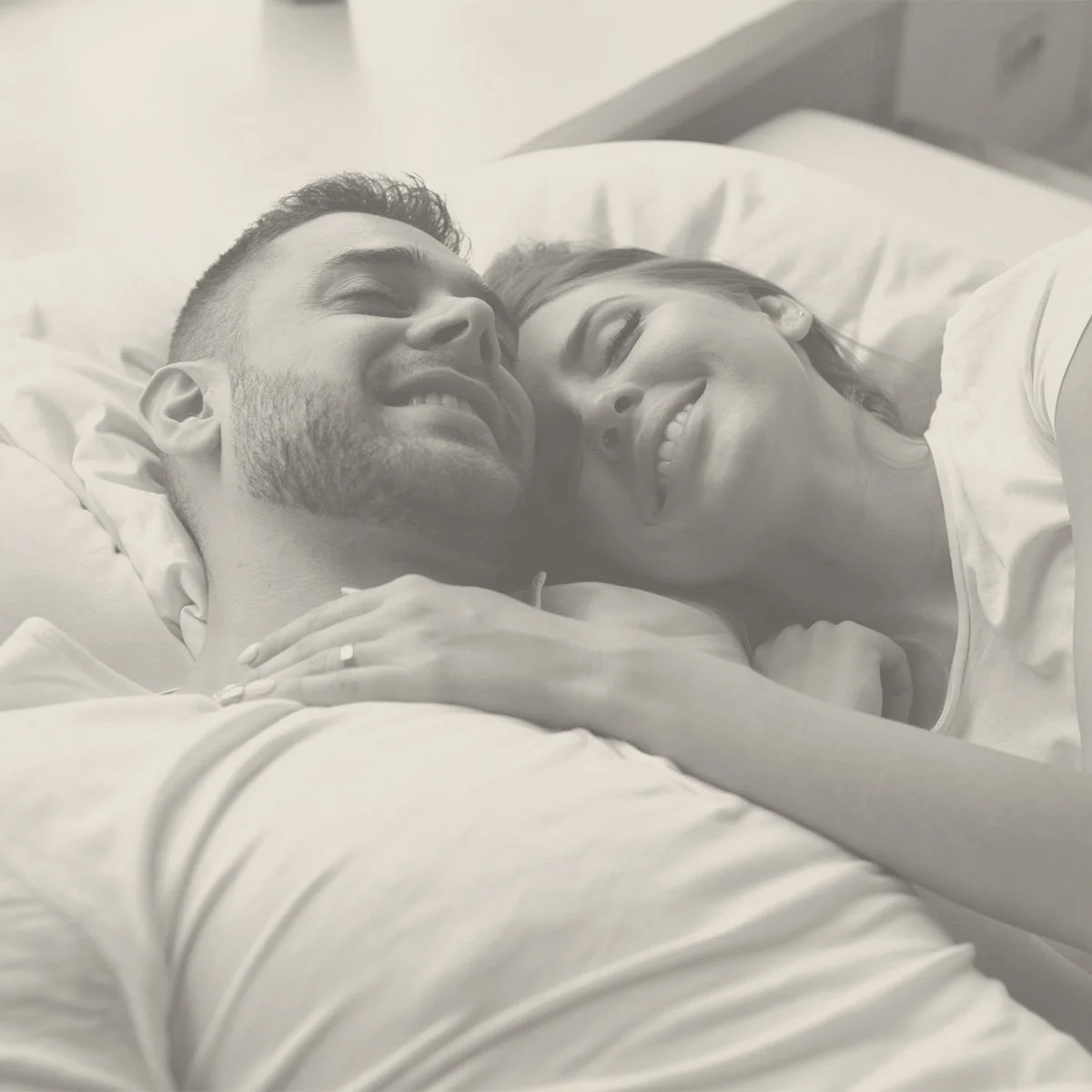 A smiling couple lying in bed, looking at each other affectionately.