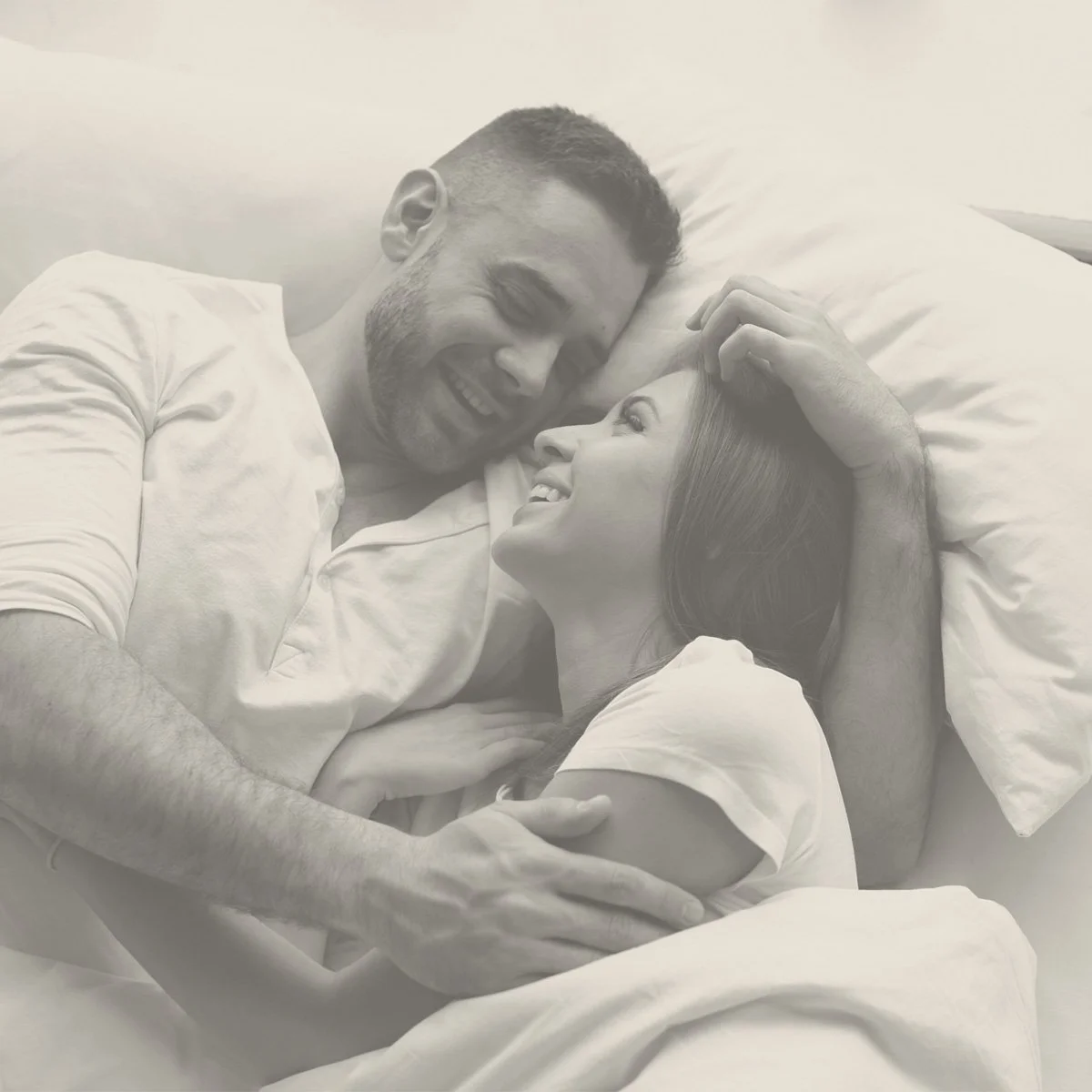 A happy couple lying in bed together, looking into each other's eyes, smiling, with the woman's hand resting on the man's arm.