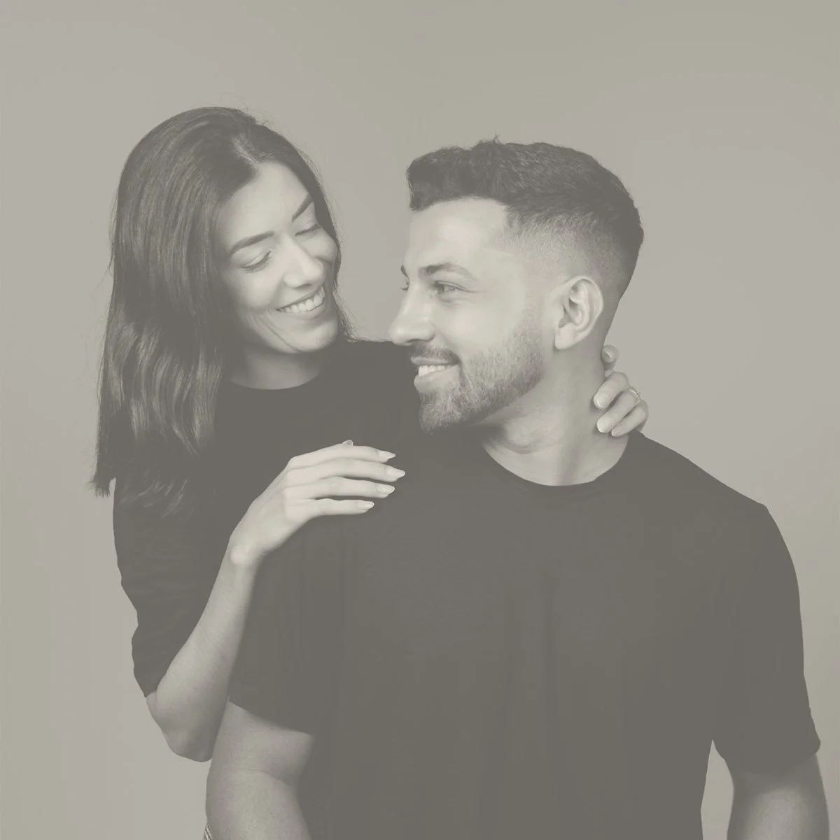 Black and white photo of a smiling woman looking at a man, who is smiling back. The woman is resting her hand on the man's shoulder and has her other hand on his neck, in a friendly, intimate pose.