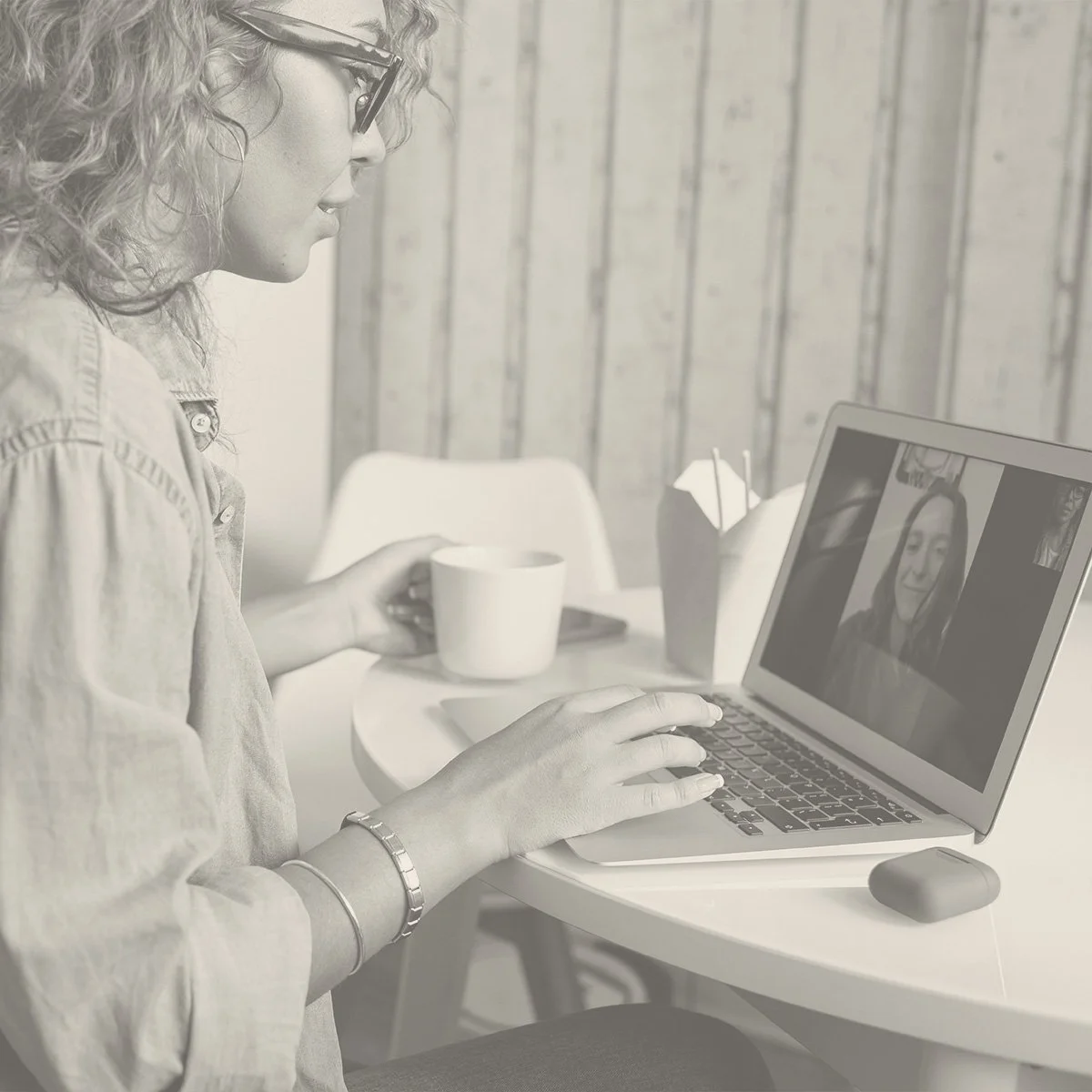 A woman with curly hair and glasses sitting at a table, engaged in a video call on her laptop. She is wearing a light-colored shirt and has a bracelet on her wrist. On the table, there is a coffee cup, a box of tissues, and a mouse. The background features a wooden wall.